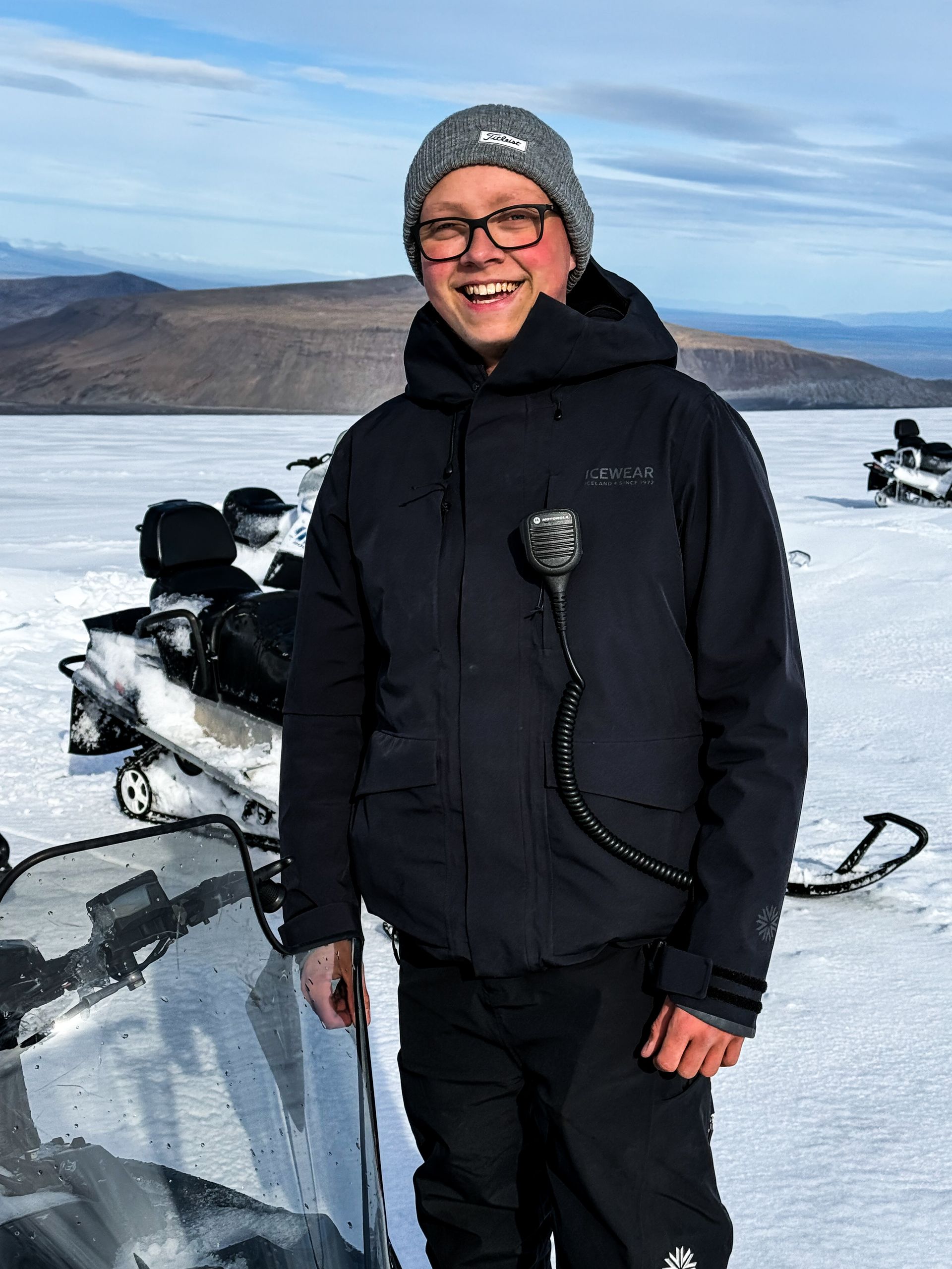 Man in winter jacket and hat, standing by snowmobiles on snow-covered terrain. Smiling, with a microphone.