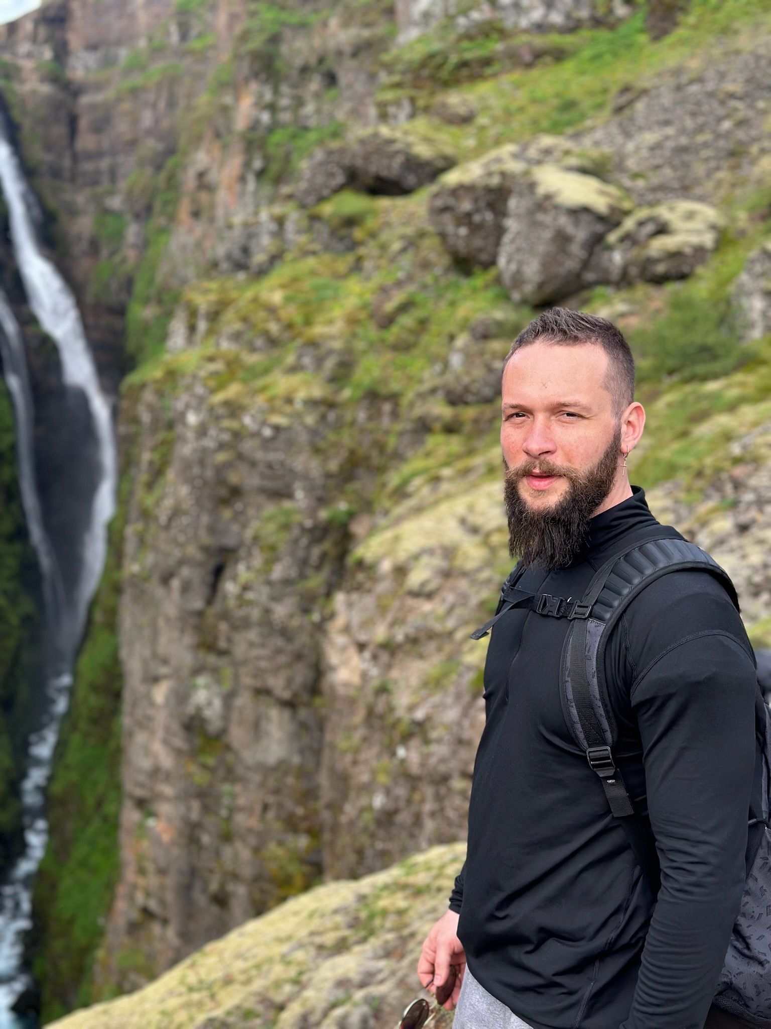 Man with beard, black shirt, backpack, by waterfall. Green moss, rocks in background.