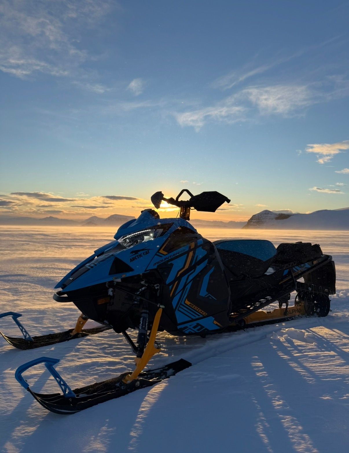 Blue snowmobile on snowy expanse, backlit by setting sun.
