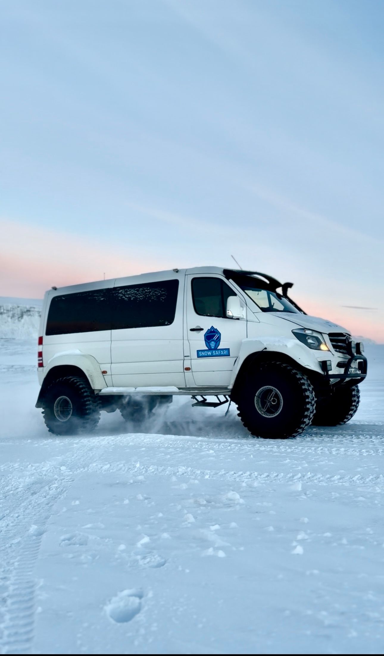 White, off-road van driving in snow. Has large tires and blue logo. Winter setting.