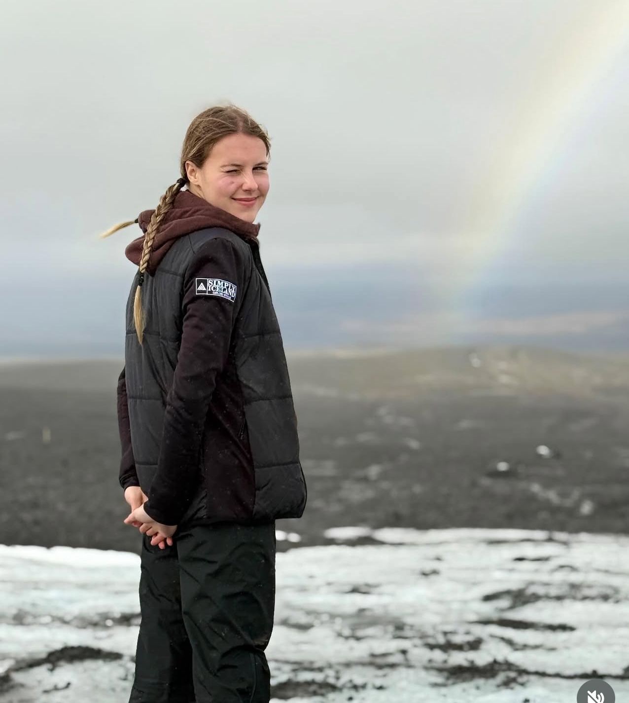 Woman squatting outdoors, smiling, with hands on cheeks. Black jacket, light setting, misty background.