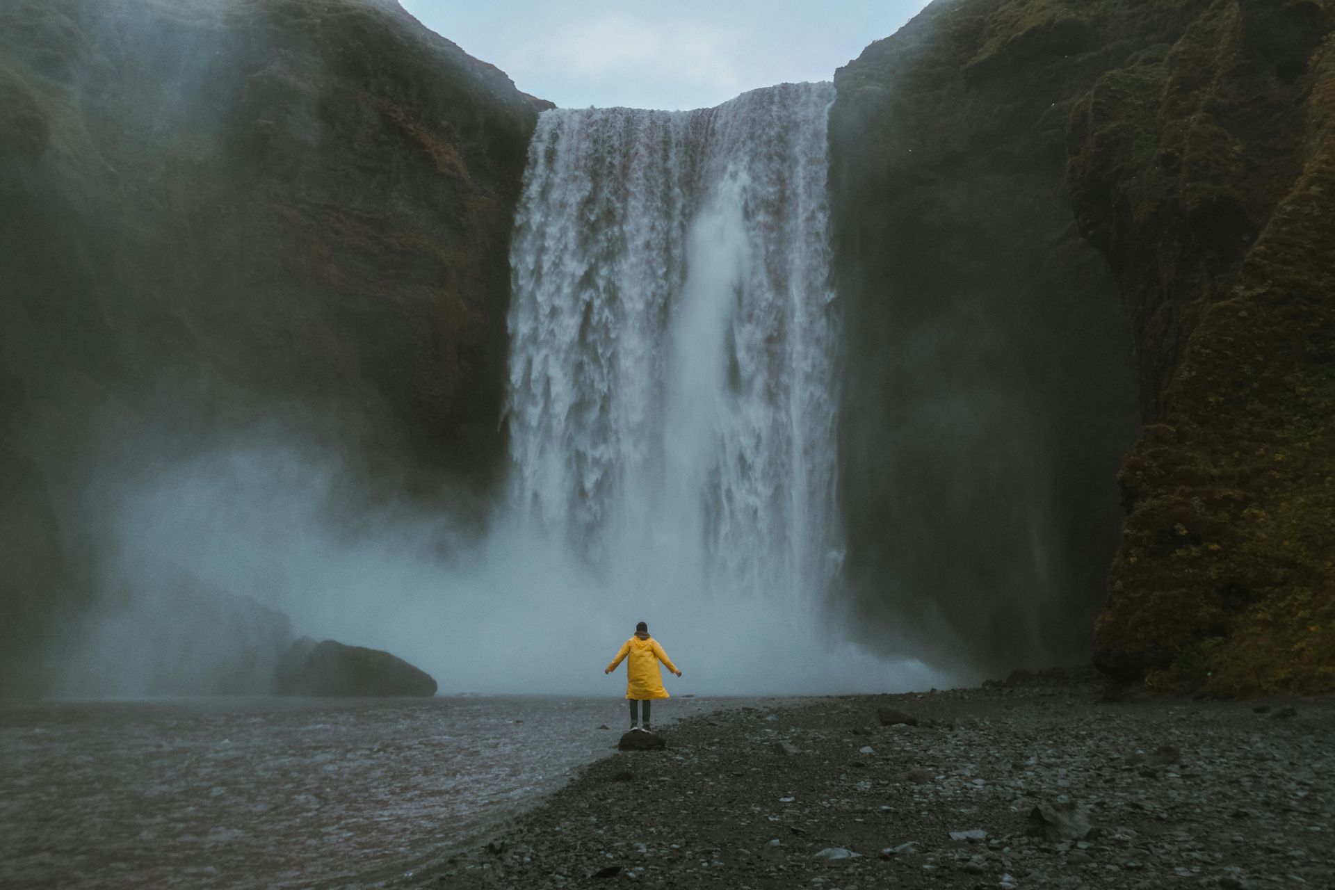 Person in yellow raincoat stands before a large waterfall, misty air, gray cliffs.
