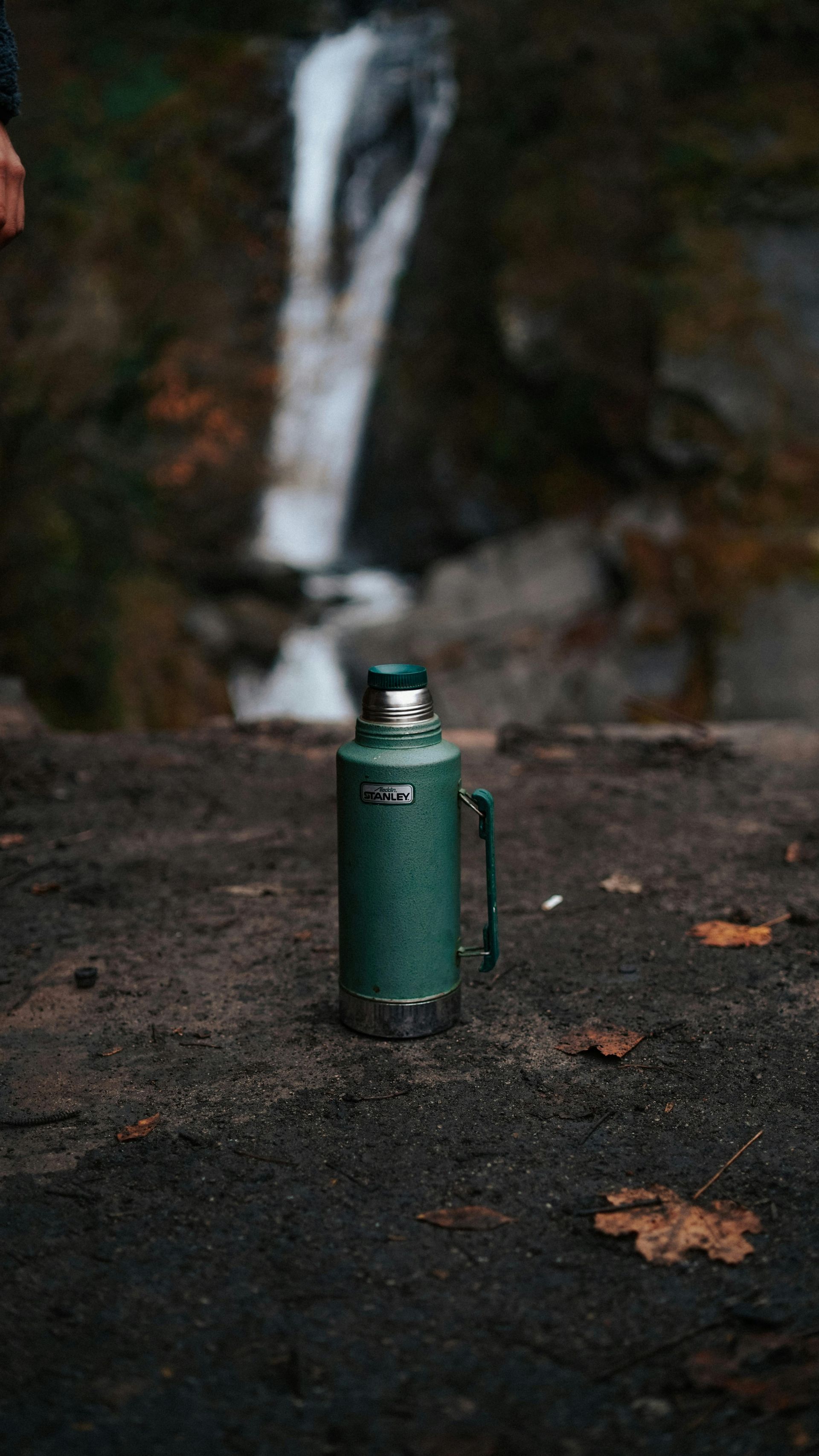 Green Stanley thermos with handle on a rock, waterfall in the blurry background.