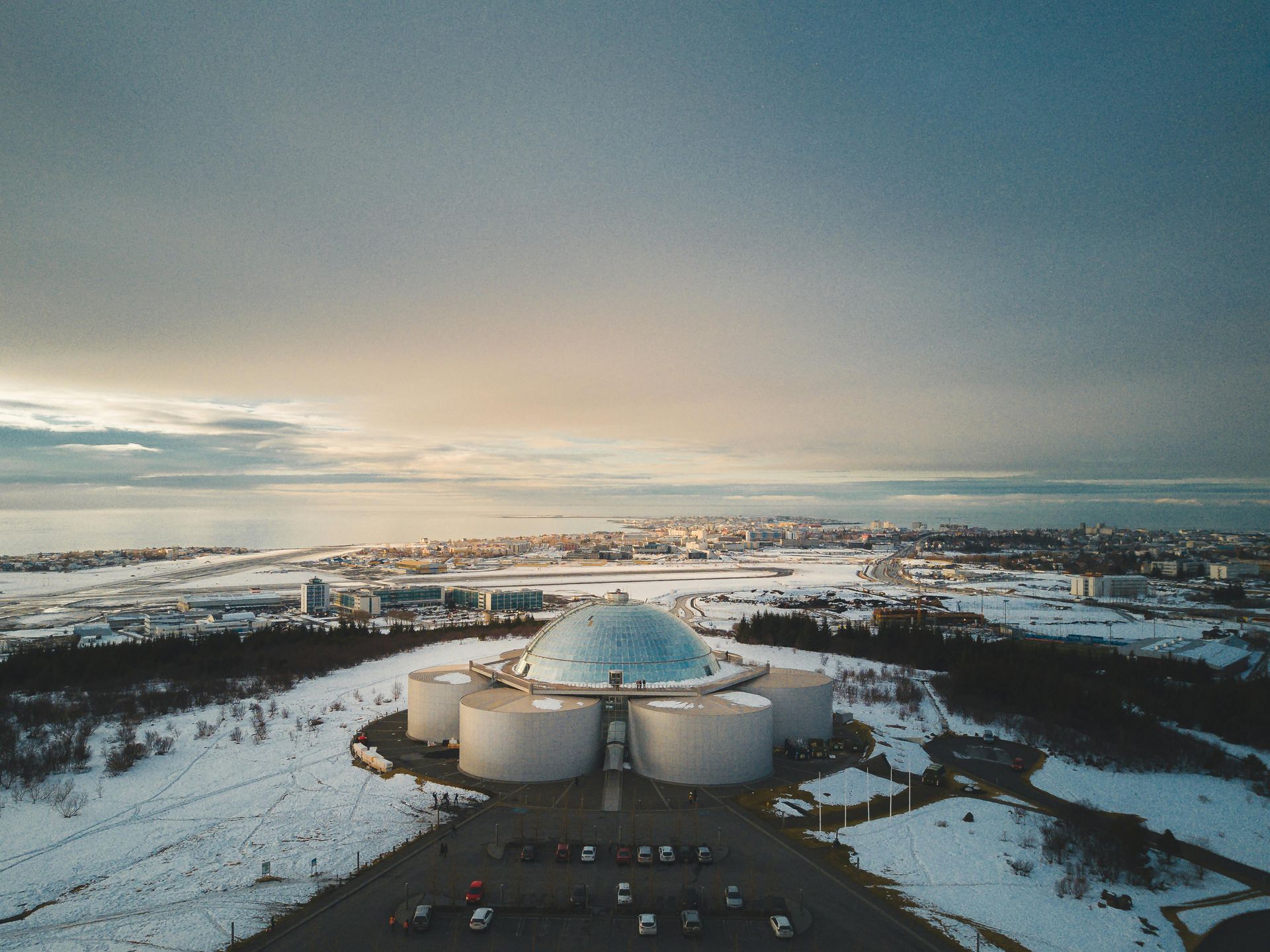 Aerial view of a large dome and cylindrical tanks on a snow-covered hill overlooking a city and ocean under a cloudy sky.