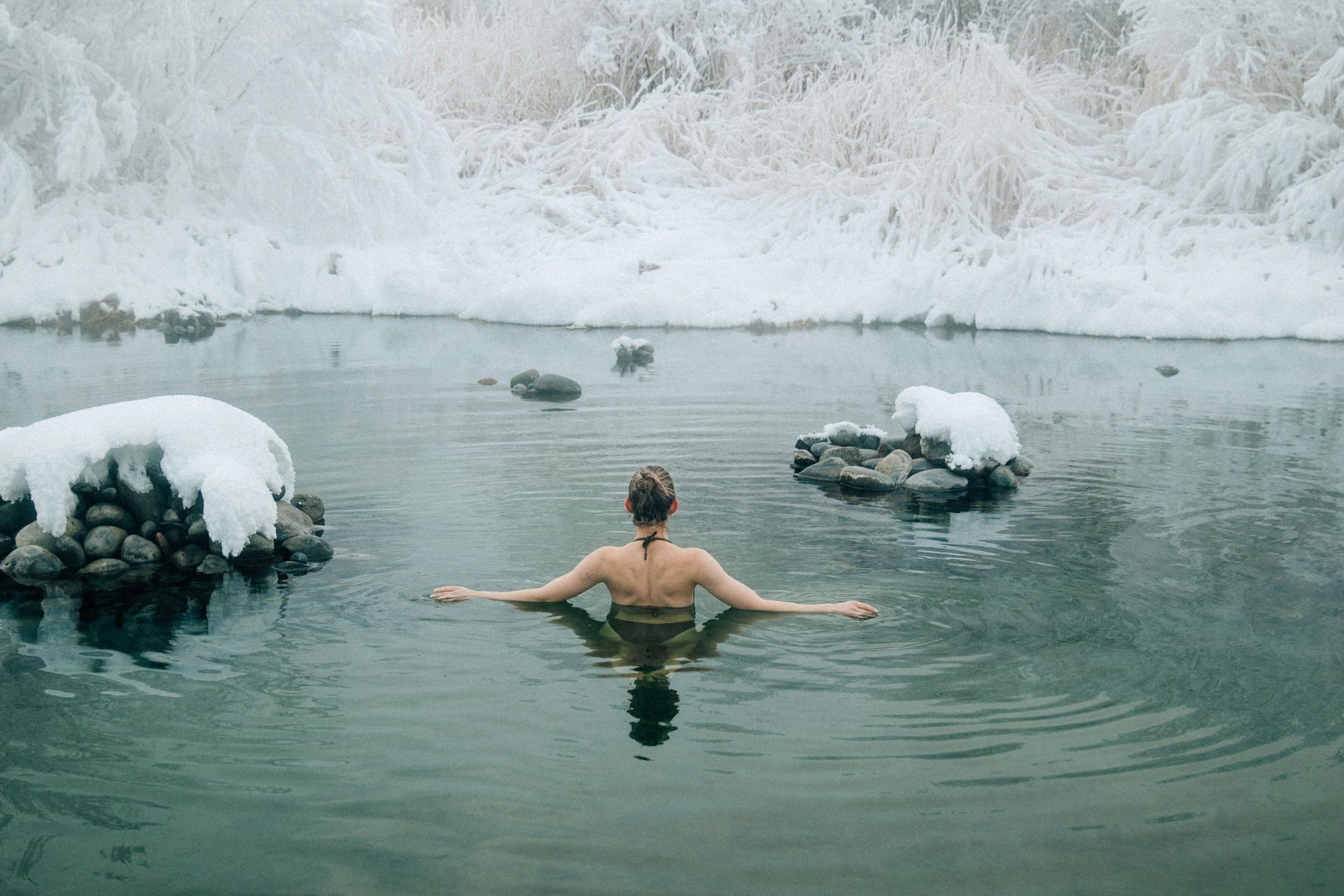 Woman in swimwear in a snow-covered pond, arms outstretched, surrounded by icy landscape.