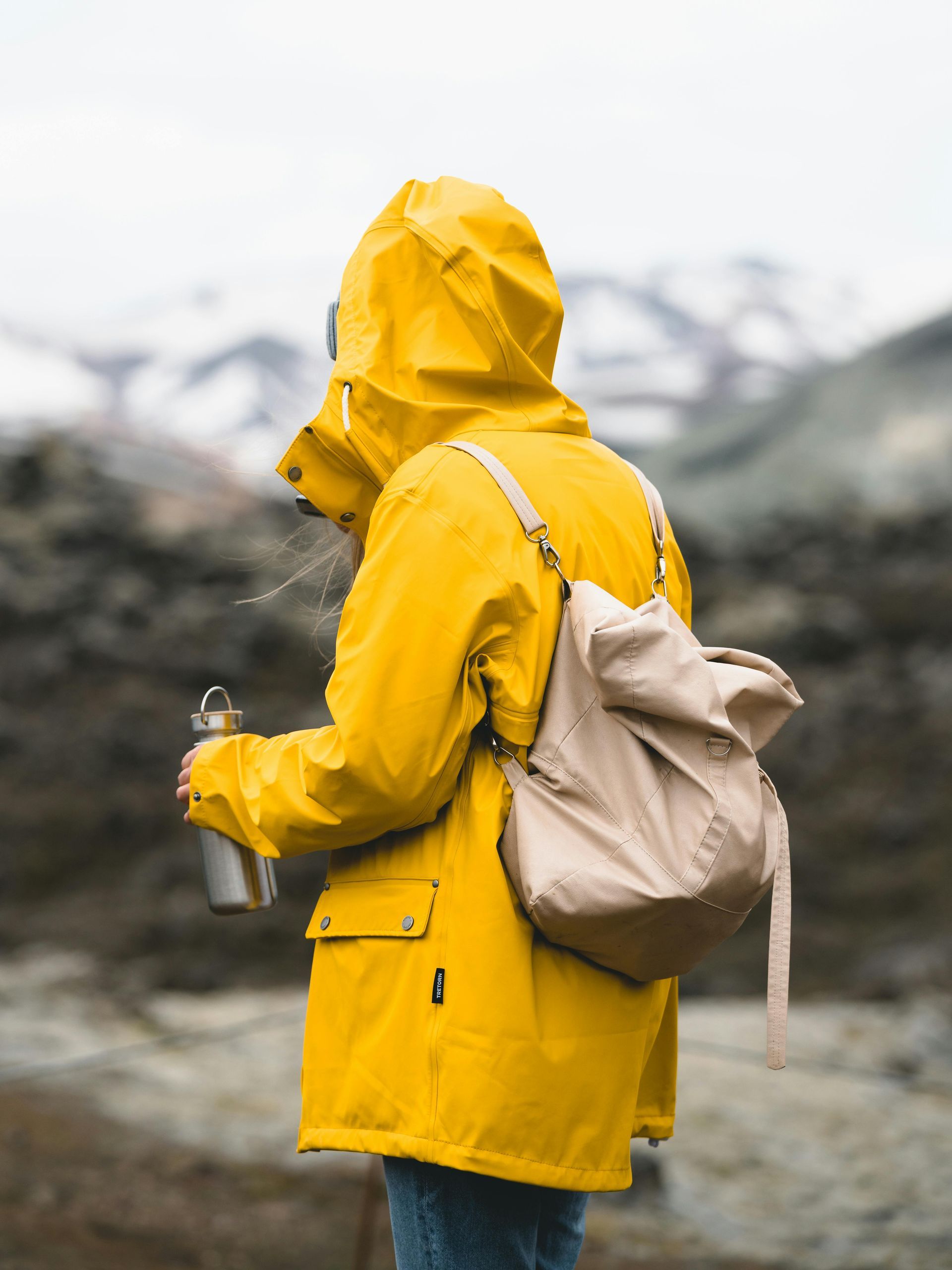 Person in yellow raincoat, tan backpack, holding a metal water bottle, standing outdoors with a blurred mountain background.