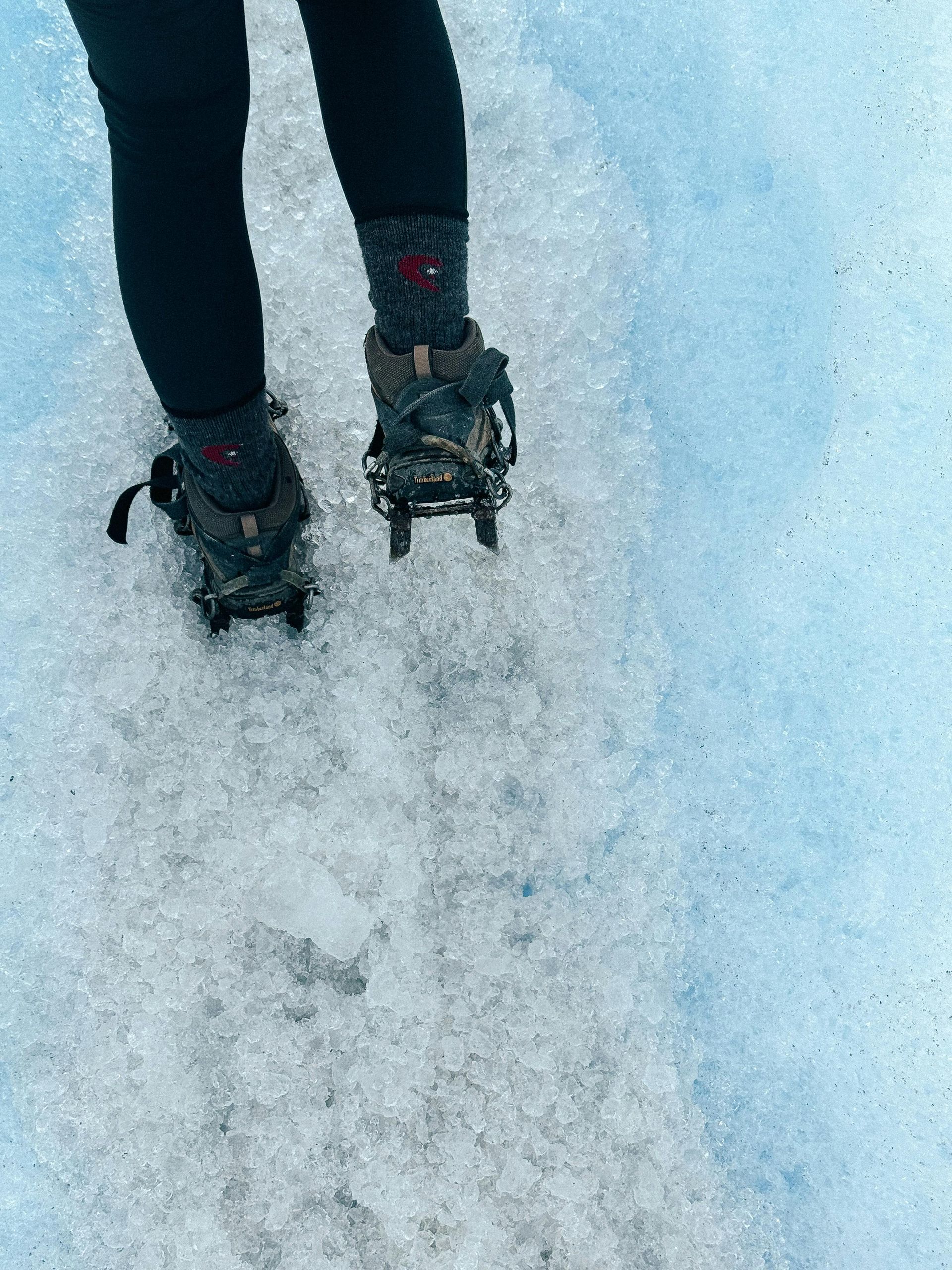Person's legs in black pants and boots with crampons walking on a blue glacier.