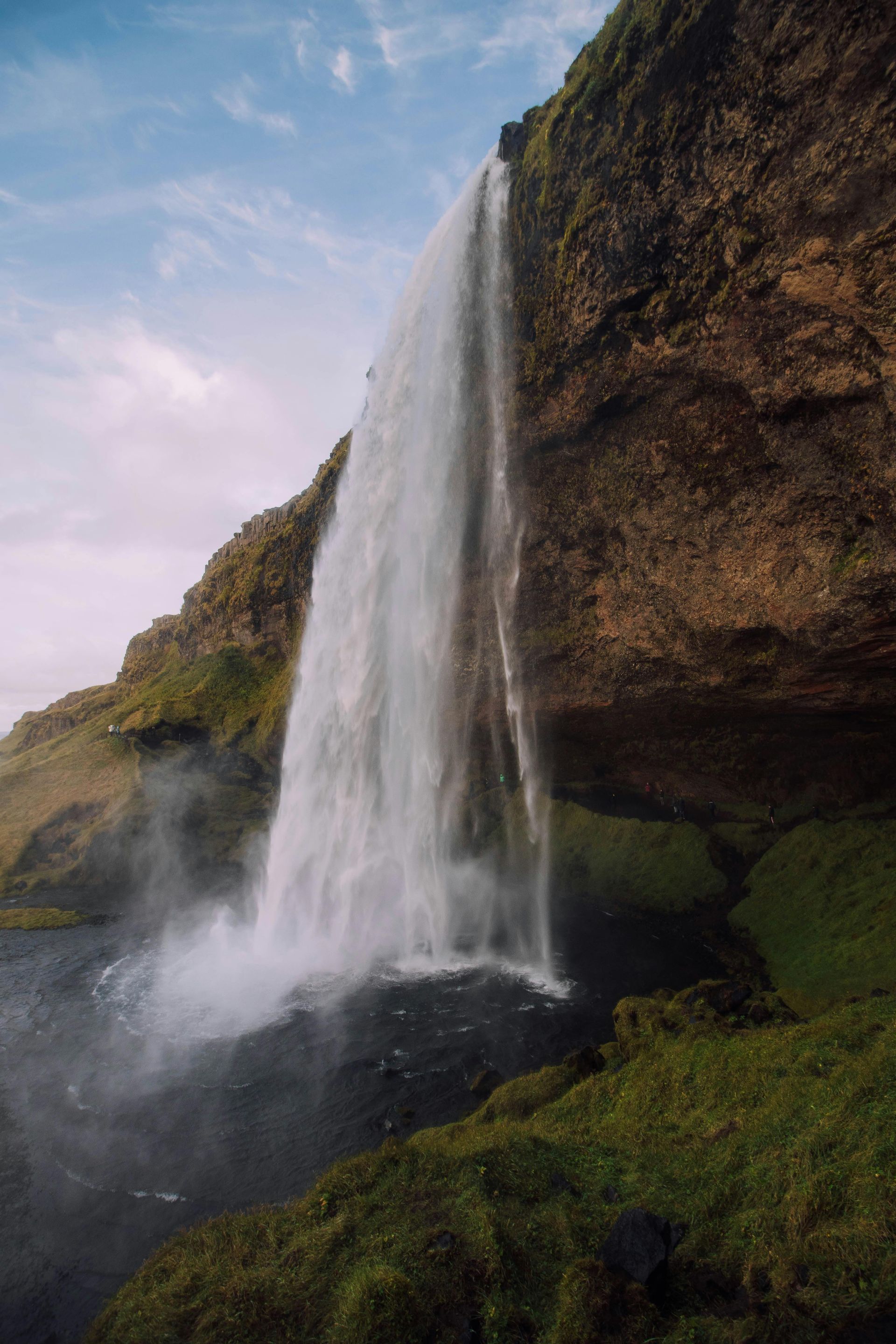 Waterfall cascading down a cliffside, mist rising, surrounded by green grass and overcast sky.