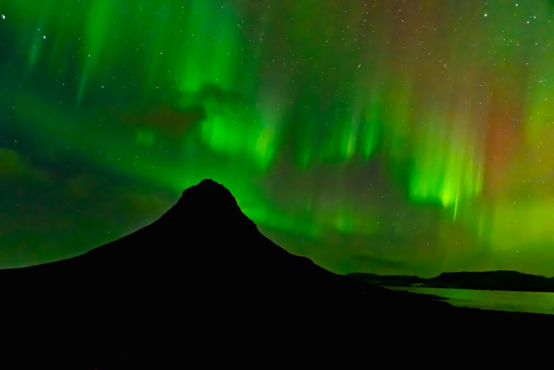 Green and red aurora borealis over a silhouetted mountain and water at night.