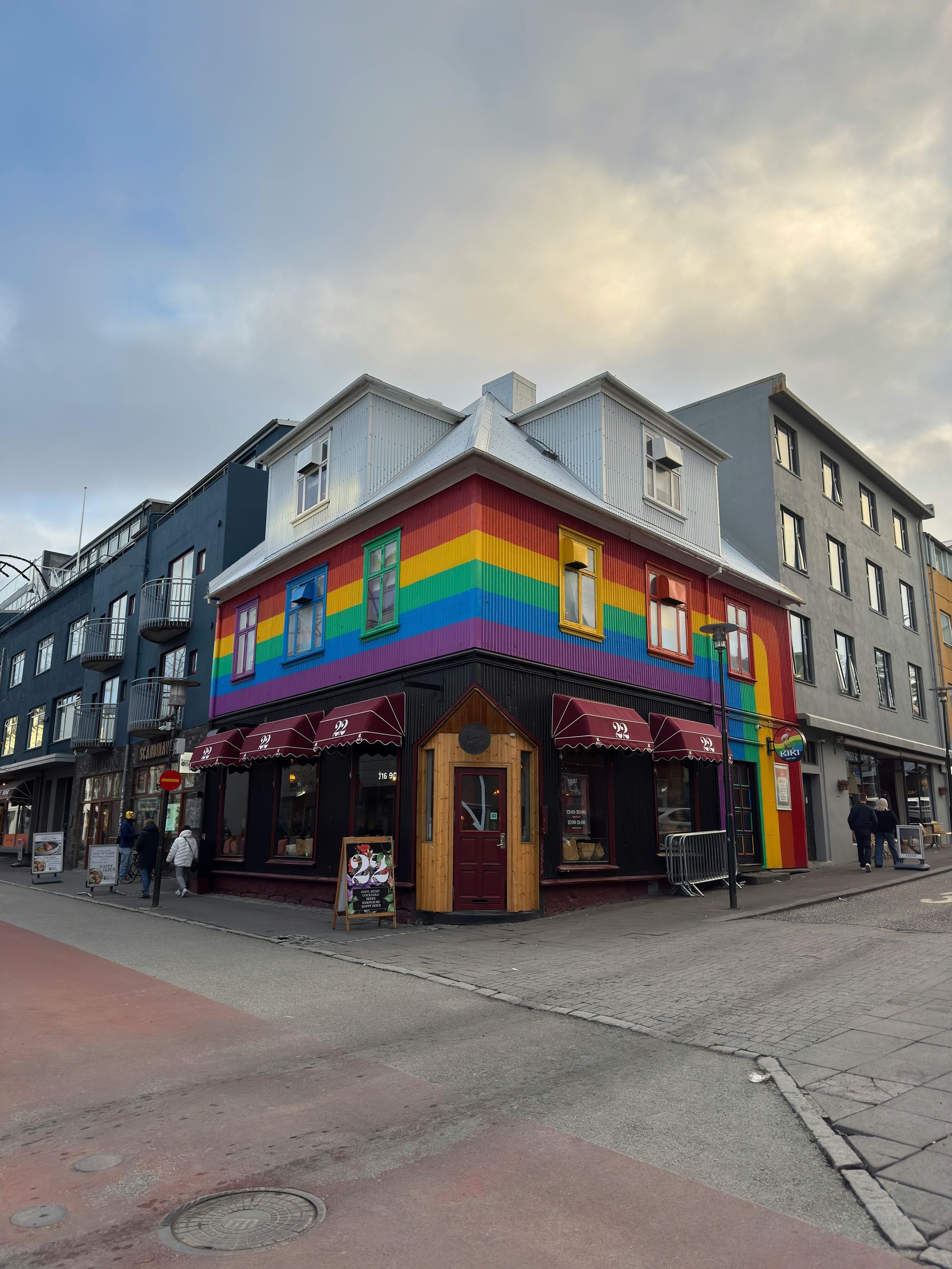 Rainbow-painted building on a corner, with red awnings. A street with people and other buildings.
