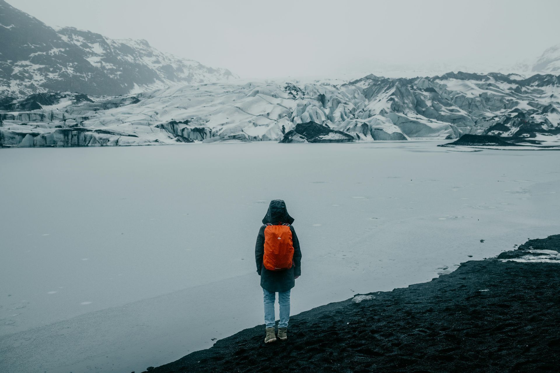 Person with an orange backpack stands before a glacial lake and snowy mountains on an overcast day.