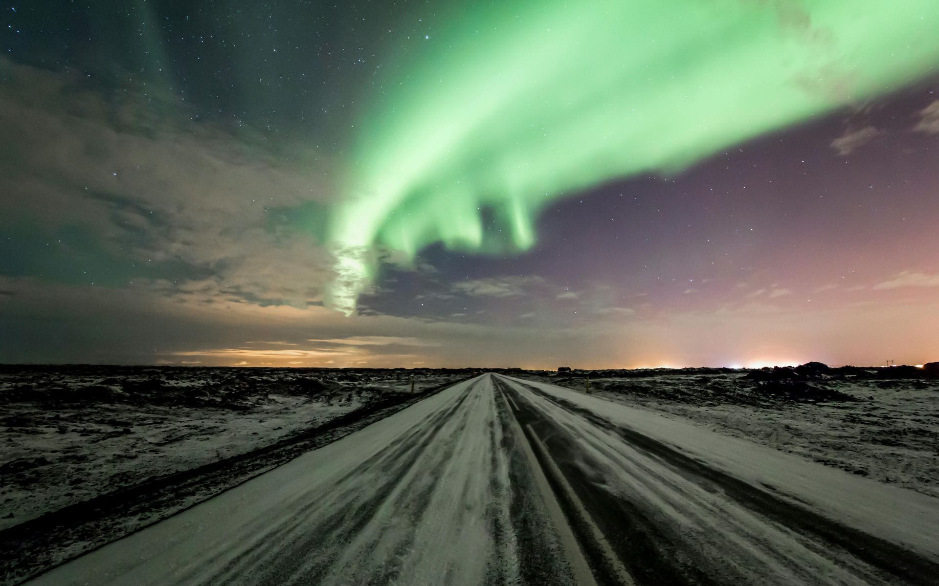 Green aurora borealis streaks across a starry sky above a snow-covered road.