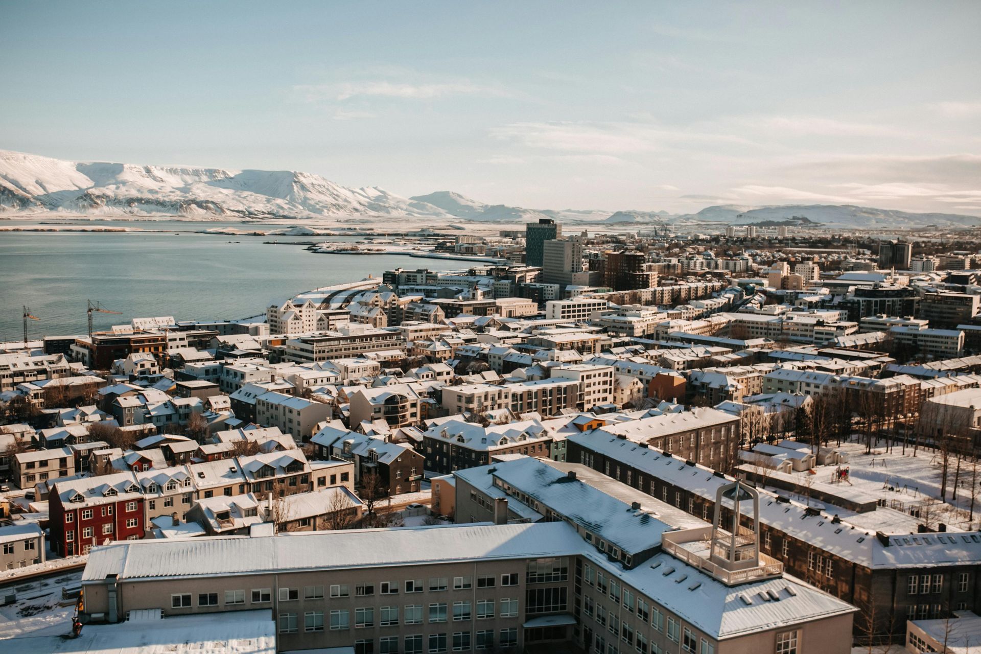 Snowy city view with ocean and mountains in the distance under a blue sky.
