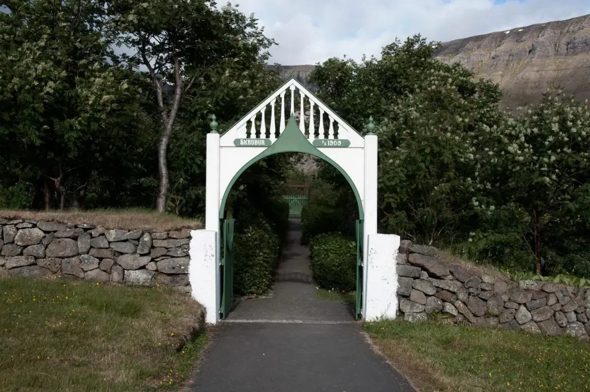 White arched gate entrance to a garden pathway, stone wall, green foliage, mountain backdrop.