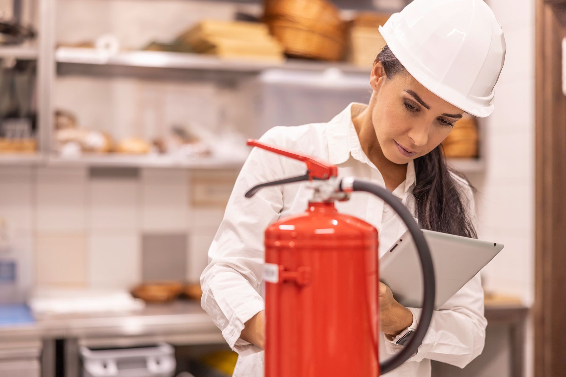 Woman in hard hat inspecting a red fire extinguisher, holding a tablet in a kitchen.Imag