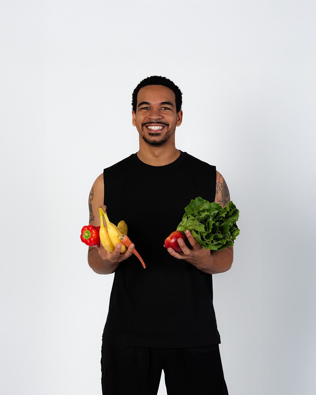 A man in a black tank top is holding fruits and vegetables in his hands.