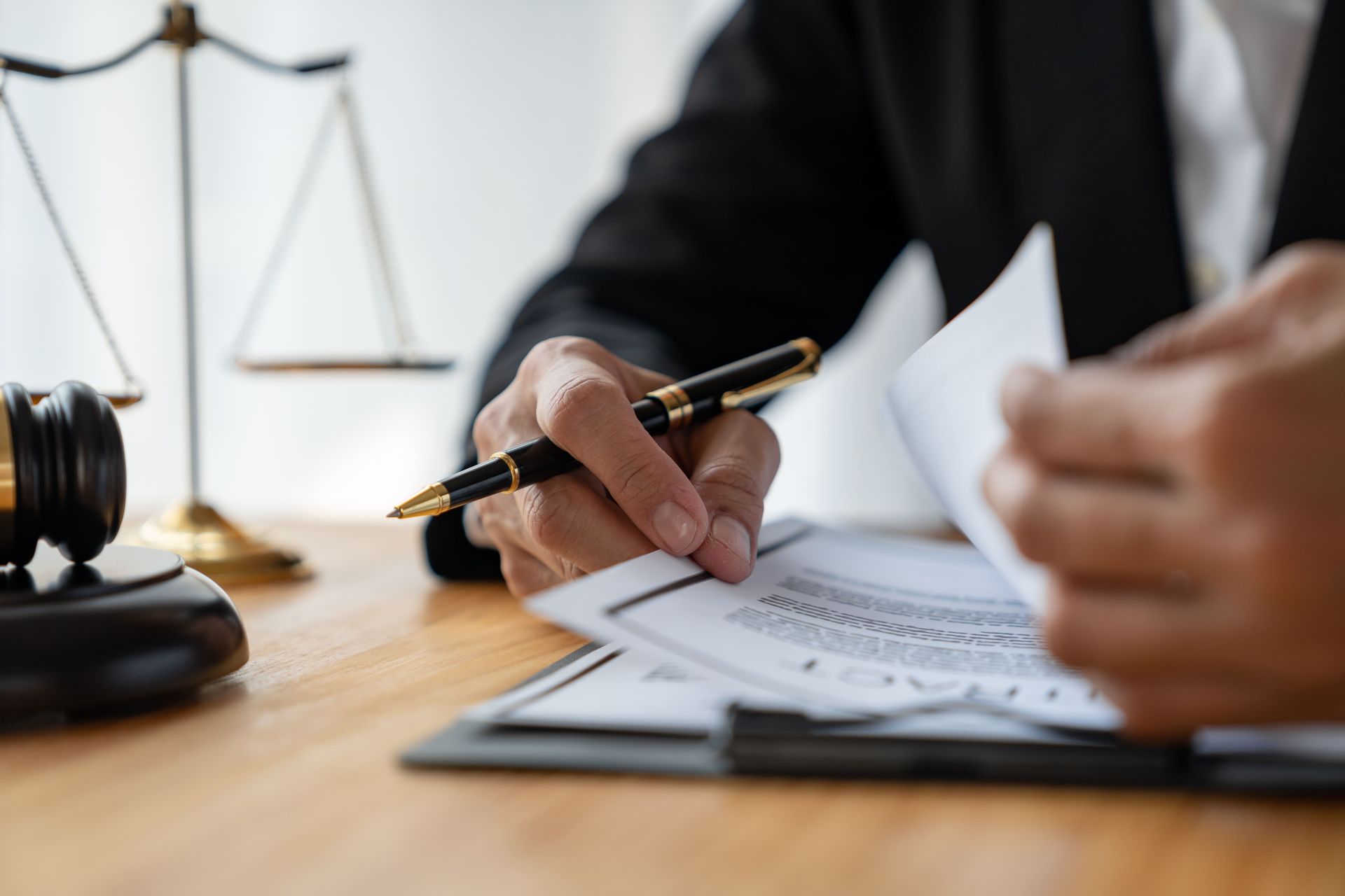 A person in a suit reviews a legal contract with a fountain pen, next to a wooden gavel and scales of justice.