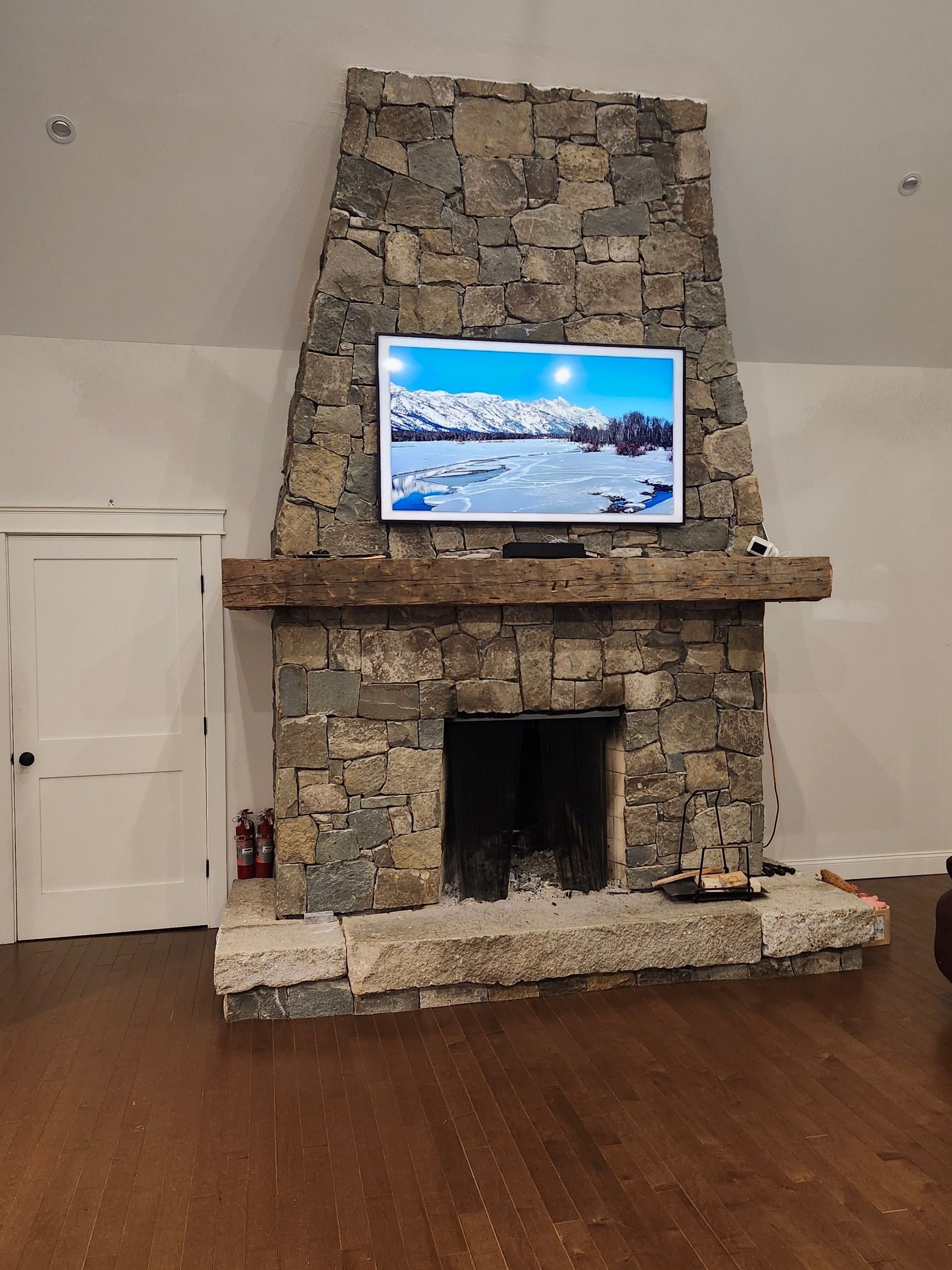 A tall, rustic stone fireplace with a thick wooden mantle and a mounted television displaying a snowy mountain scene.
