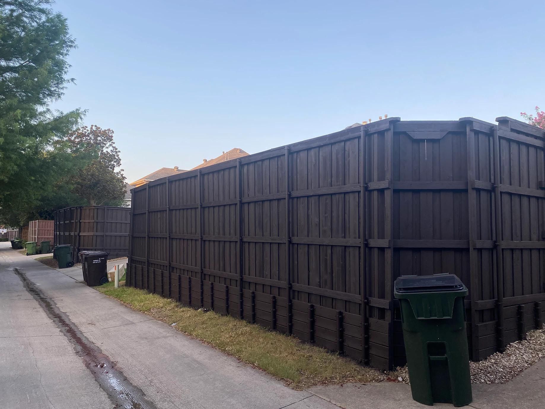 A wooden fence is surrounded by trash cans on the side of the road.