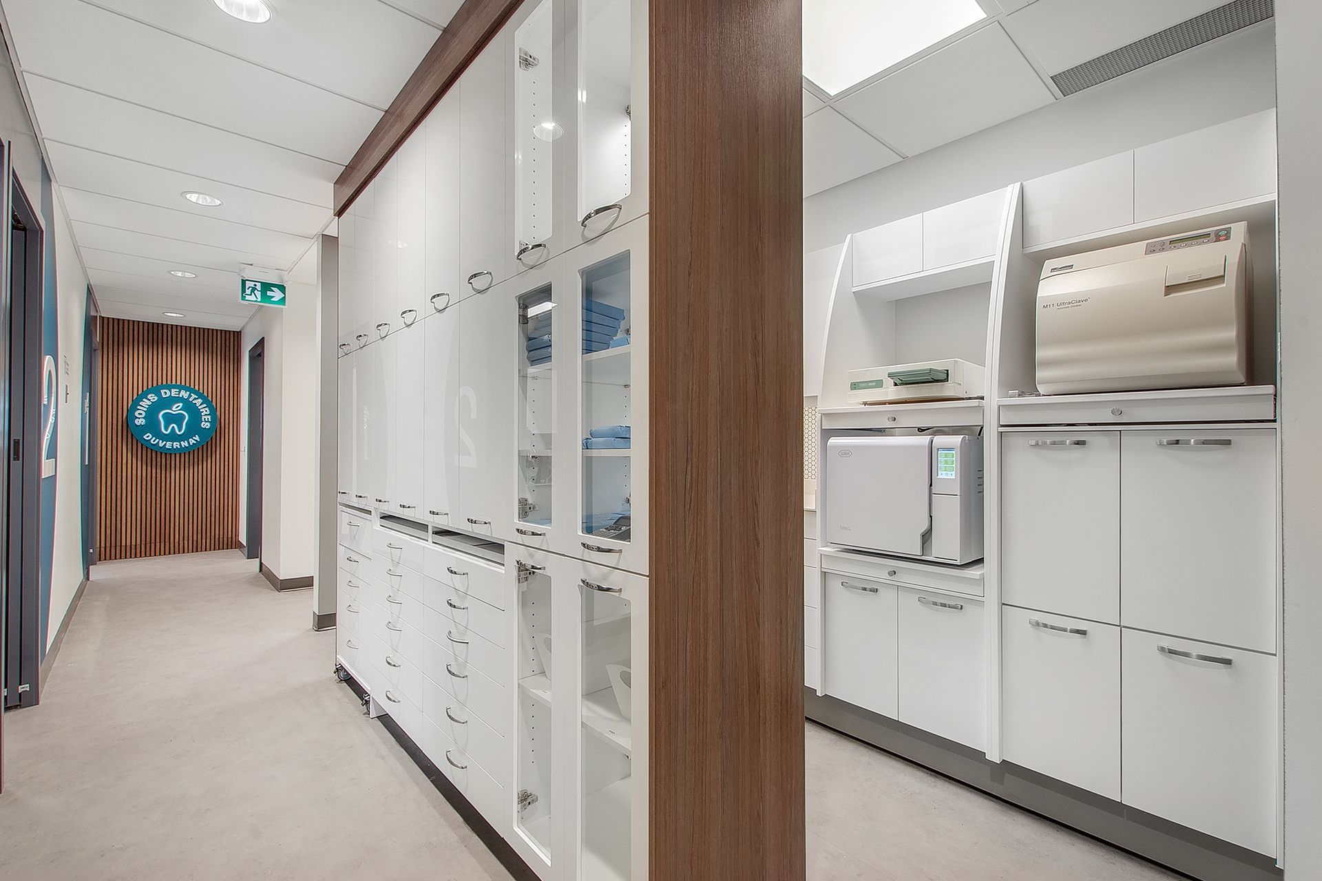 Une salle de stérilisation dentaire stérile avec des armoires et du matériel blancs, adjacente à un couloir avec un panneau circulaire turquoise.
