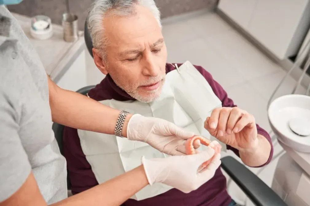 Dans un cabinet dentaire, un professionnel portant des gants présente un dentier à un patient qui peut l'examiner.