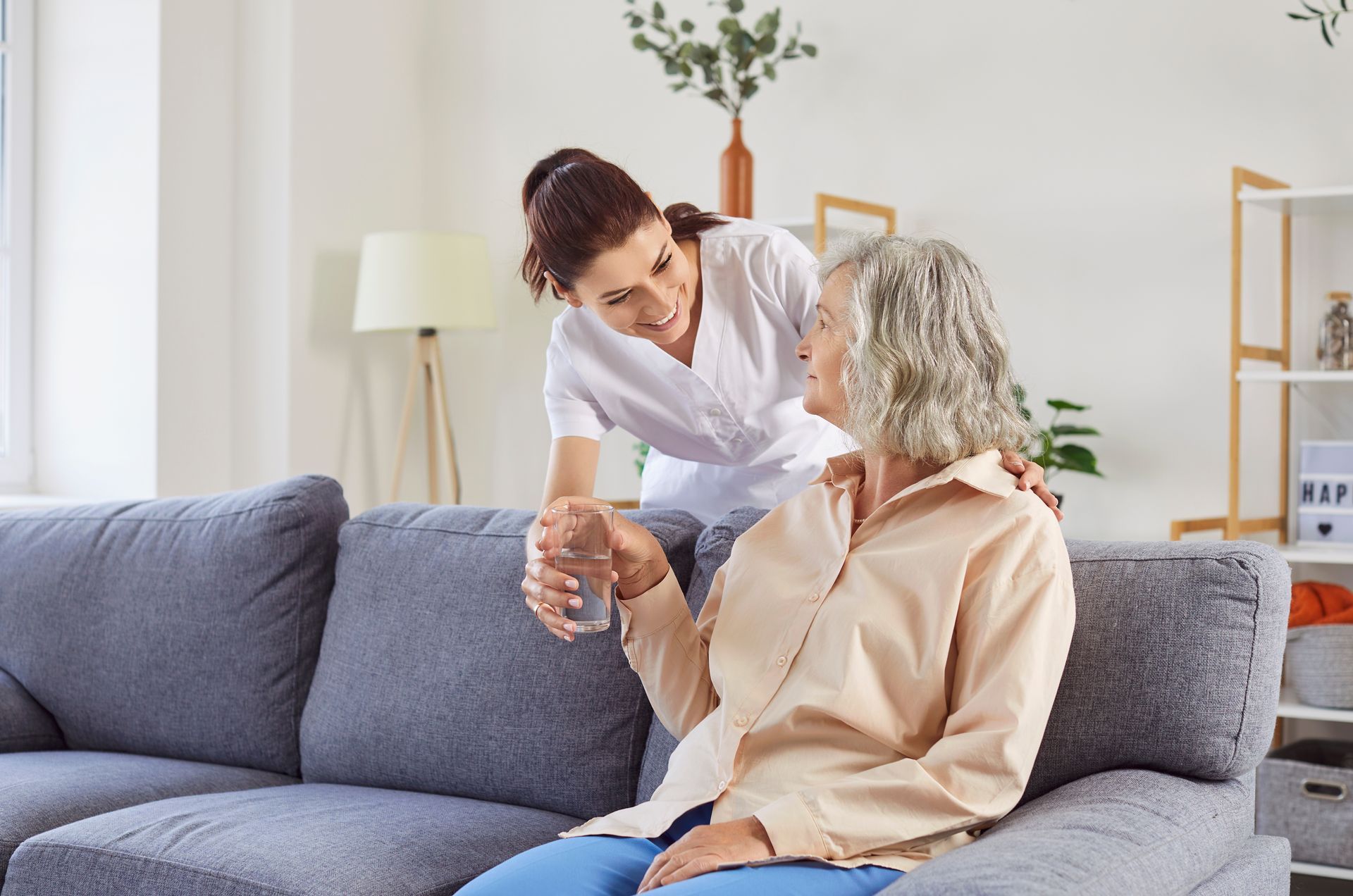 A caregiver in a white uniform hands a glass of water to an older person sitting on a grey couch in a bright living room.