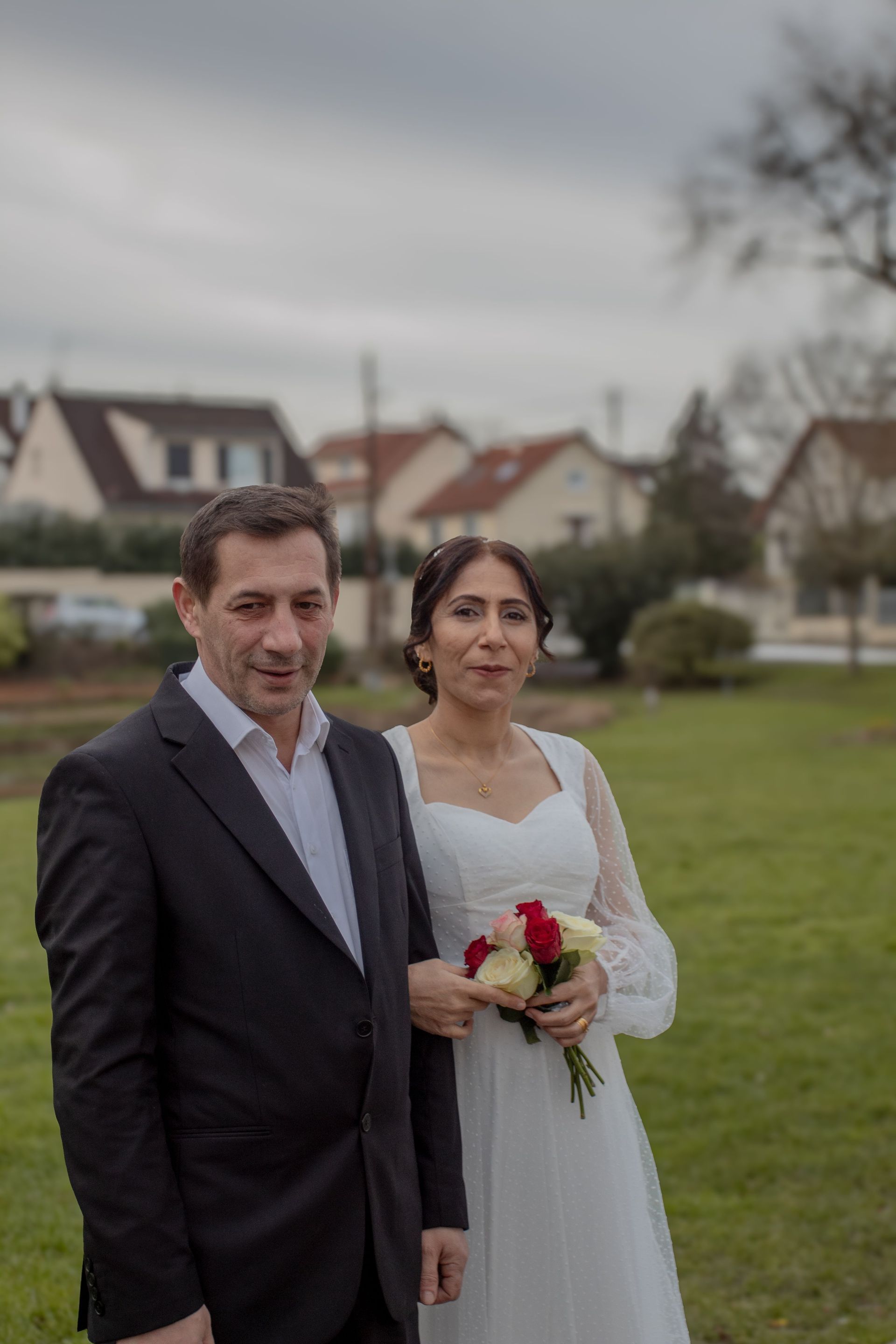 Man and bride in white dress holding a bouquet, standing on a grassy field with houses in the background.