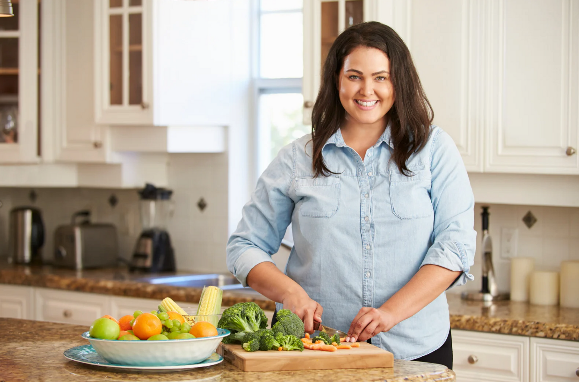 A woman is standing in a kitchen cutting vegetables on a cutting board.