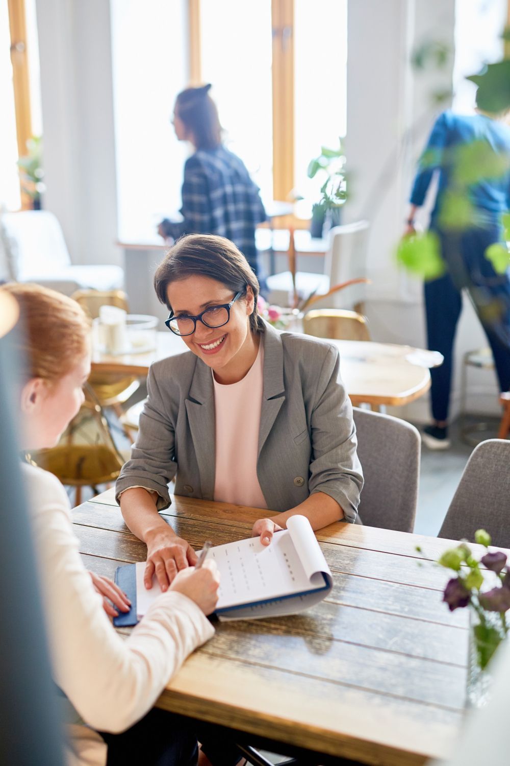 Two women reviewing a clipboard at a wooden table in a cafe. One smiles, wearing glasses and a blazer.