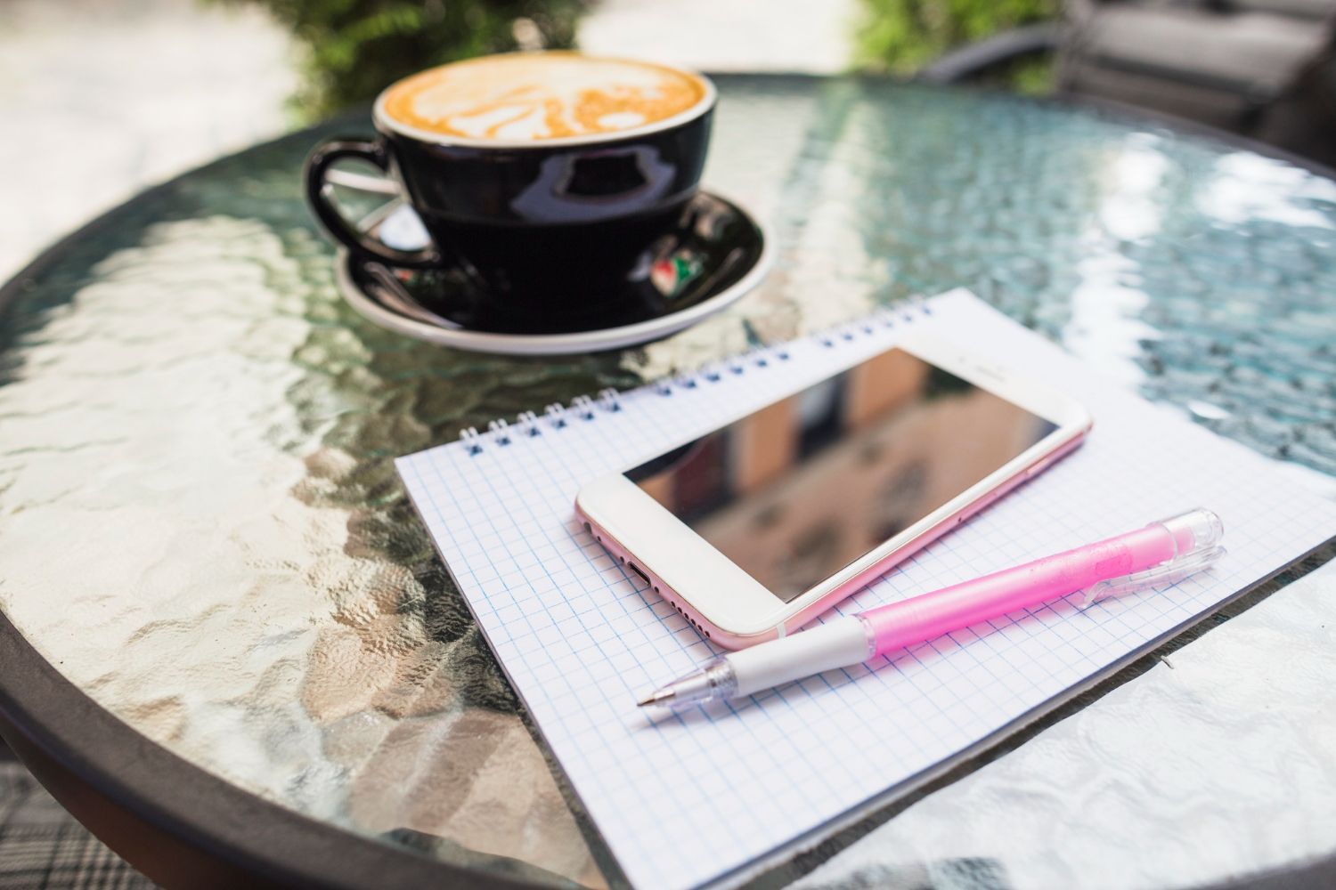 Coffee cup, notebook, smartphone, and pen on a glass table outdoors.