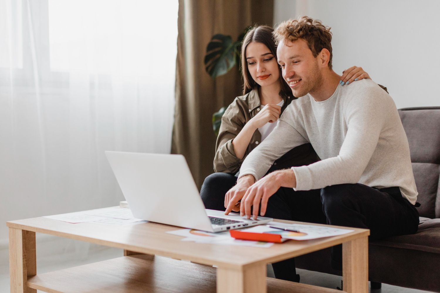 Couple on couch looking at laptop, smiling; papers and pens on table.