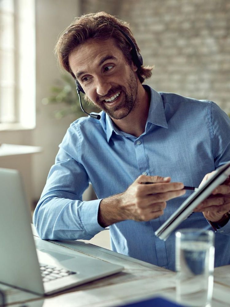 Man with headset smiles, pointing at a notepad during a video call, next to a laptop and a glass of water.