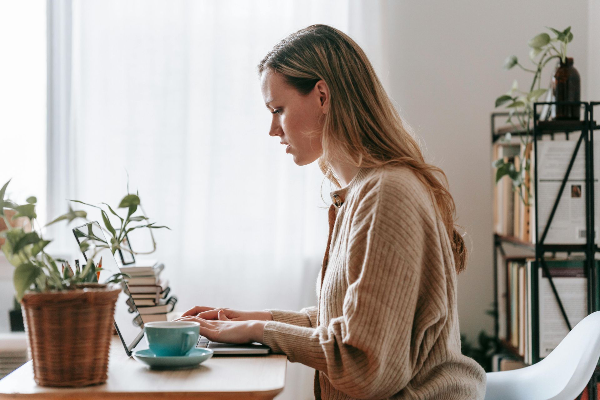 Woman with blonde hair typing on a laptop at a wooden desk, with a coffee cup, and plants near a window.