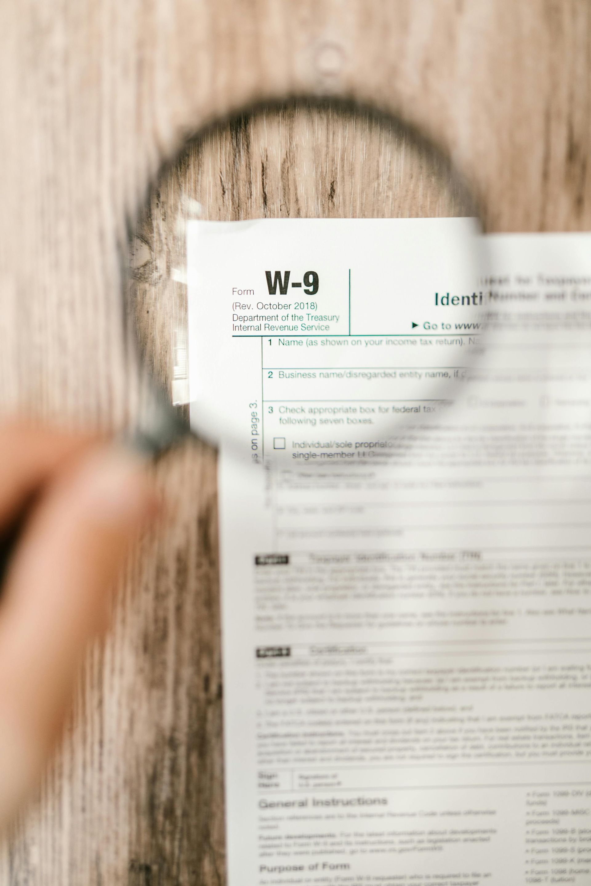Hand holding magnifying glass over a W-9 tax form on a wooden surface.