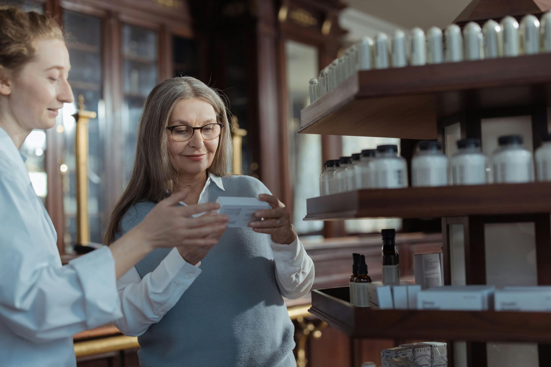 Pharmacist assisting customer, reading product box. Inside apothecary with wooden shelves, various bottles.