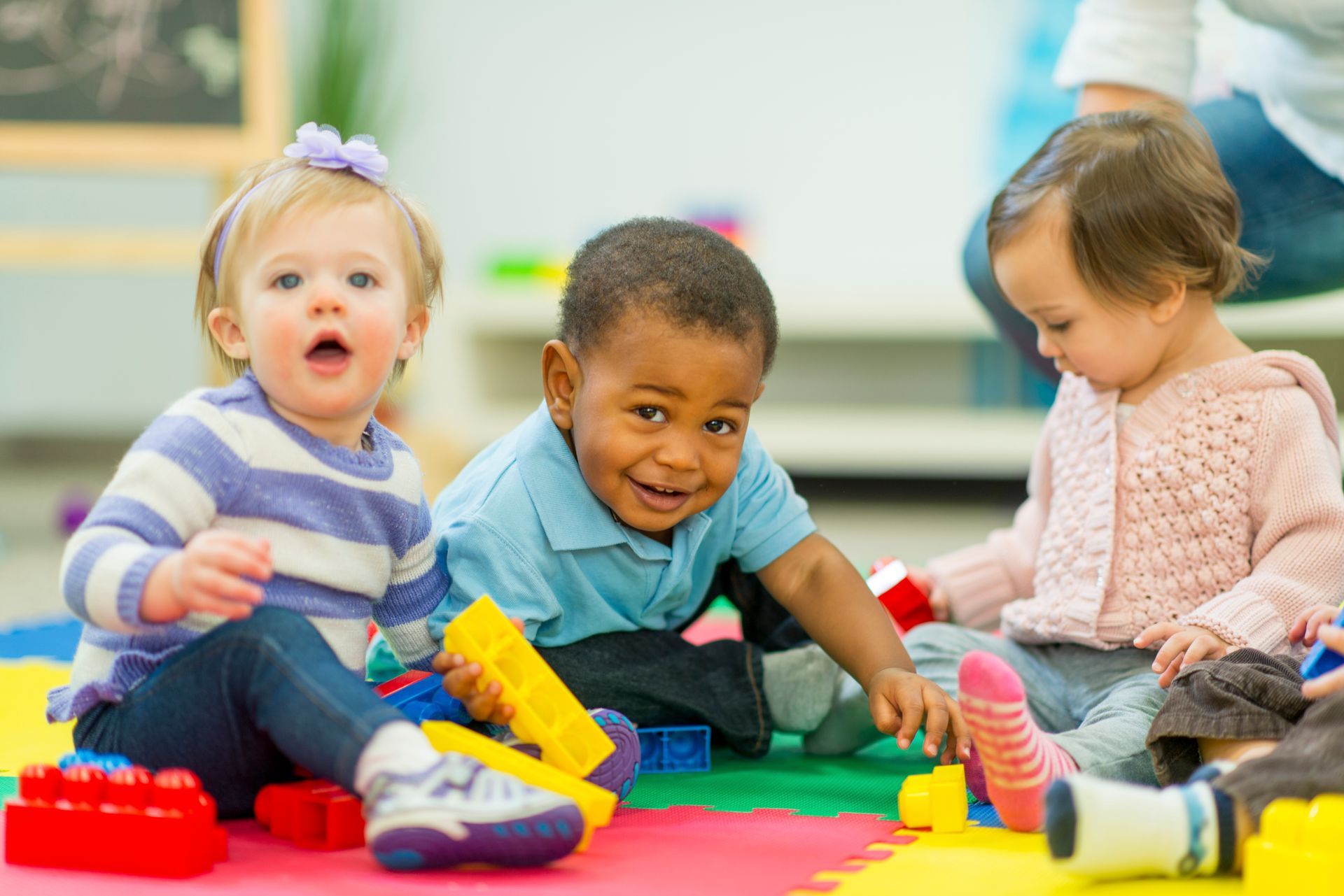 Three diverse toddlers playing with blocks on a colorful mat, one looking surprised, one smiling, and one focused.