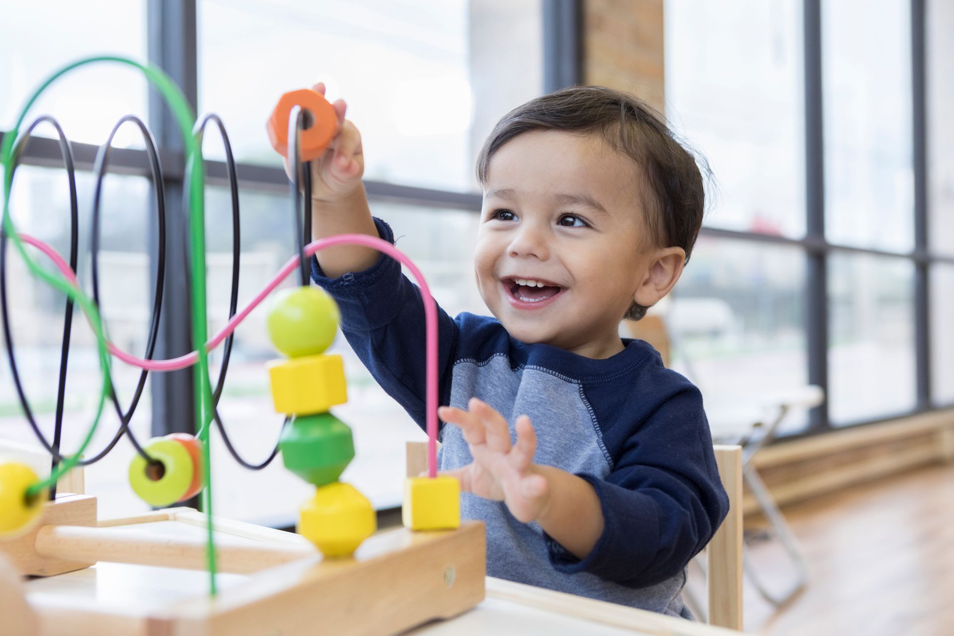 Smiling toddler playing with a wooden bead maze. He sits at a table in a room with a large window.