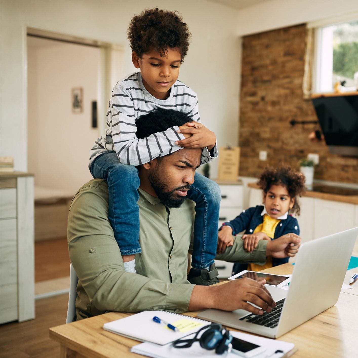 A Black man works on a laptop at a table, with a boy sitting on his shoulders and a girl reaching for him in the background. A Black man works on a laptop at a table, with a boy sitting on his shoulders and a girl reaching for him in the background.