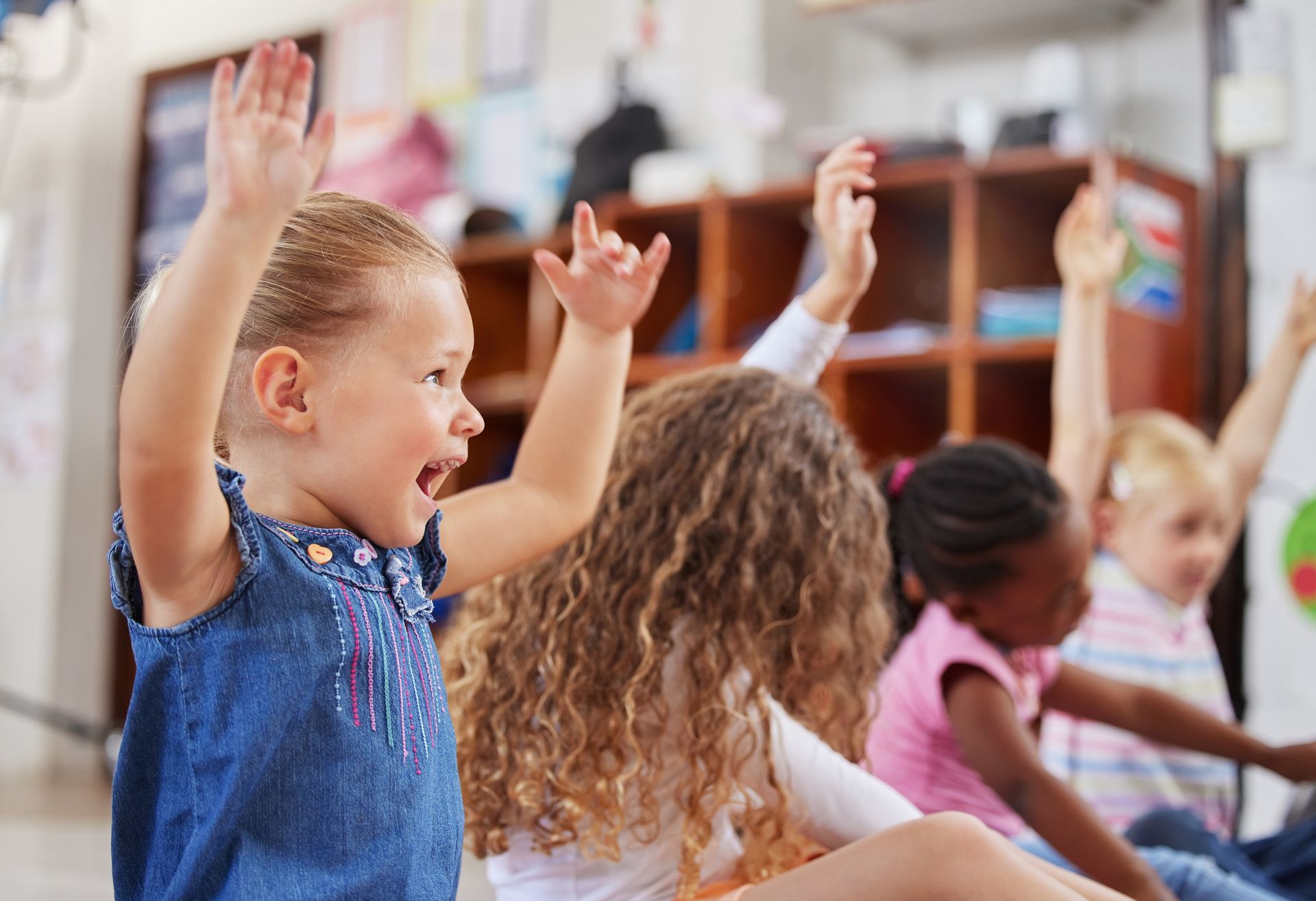 Children in a classroom raising hands. A young girl in a denim dress is excited, mouth open.