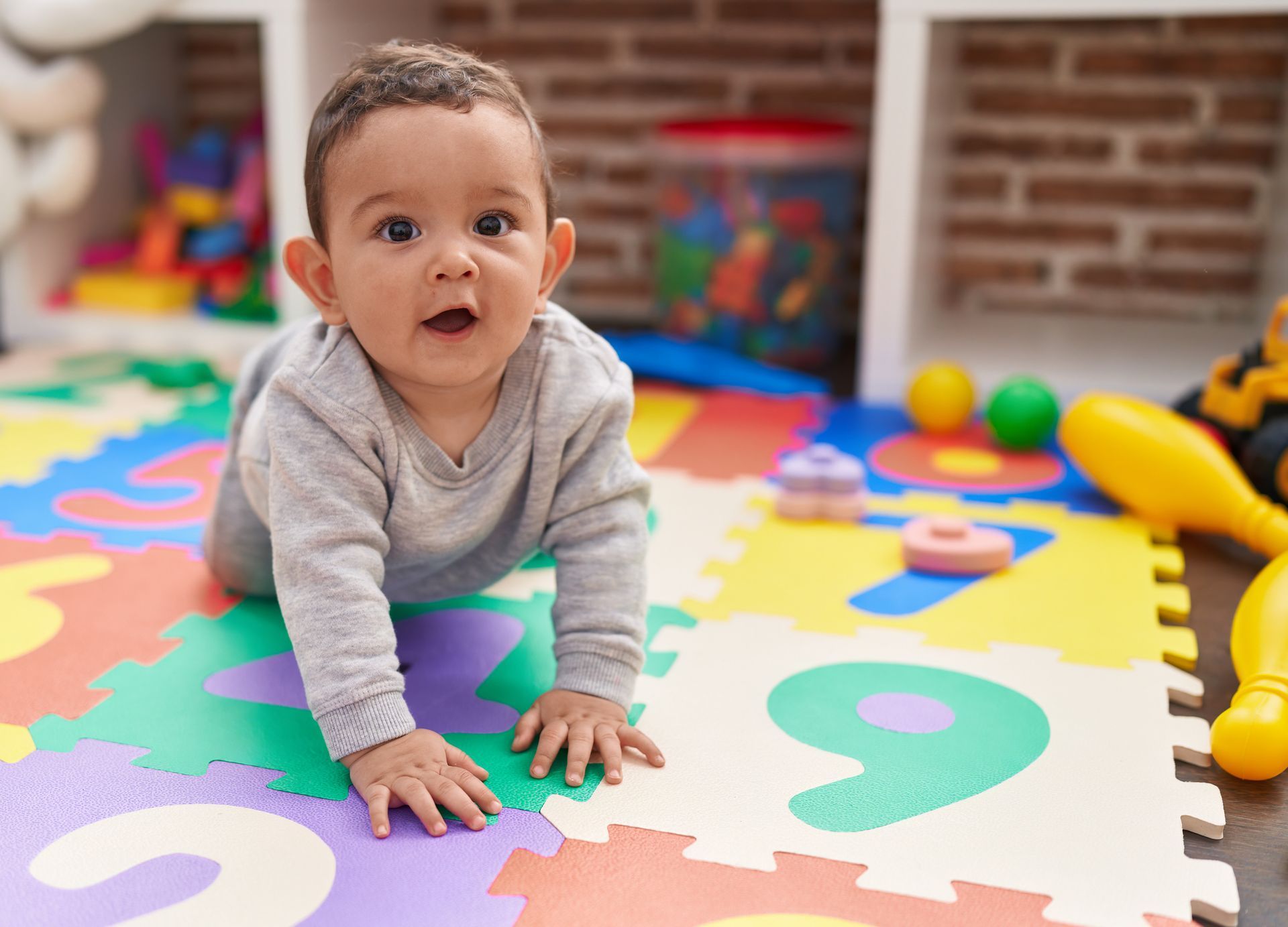Baby crawling on colorful foam playmat, looking up with surprised expression. Toys and shelves are in the background.