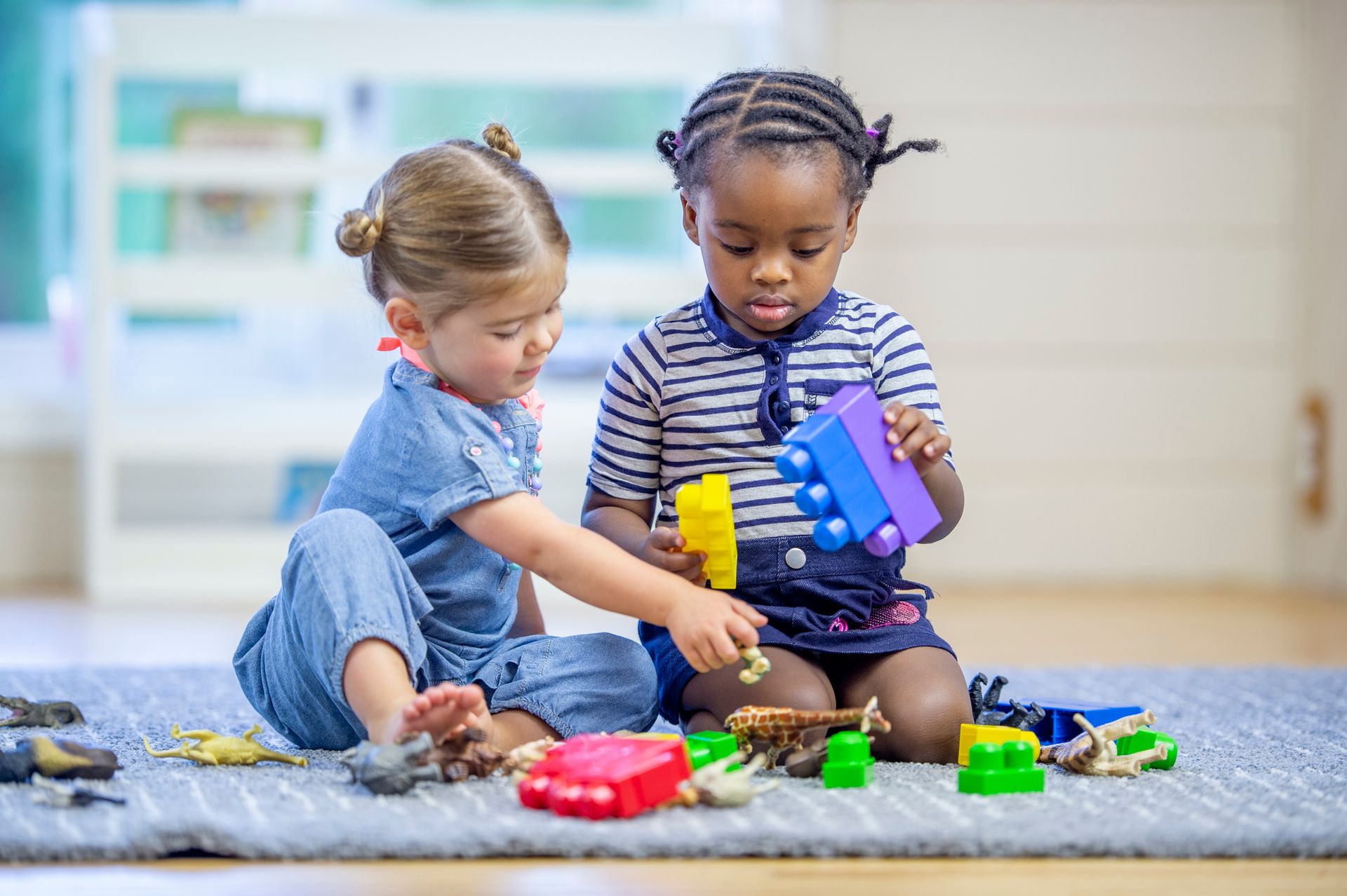 Two young girls of different races playing together on a rug, surrounded by colorful toys, indoors.