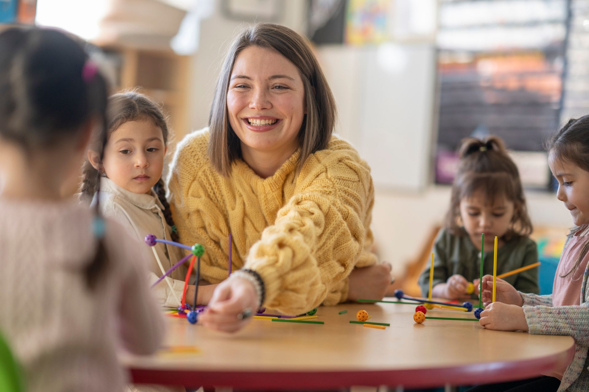 A smiling teacher in a yellow sweater helps young children assemble geometric shapes at a table in a classroom.