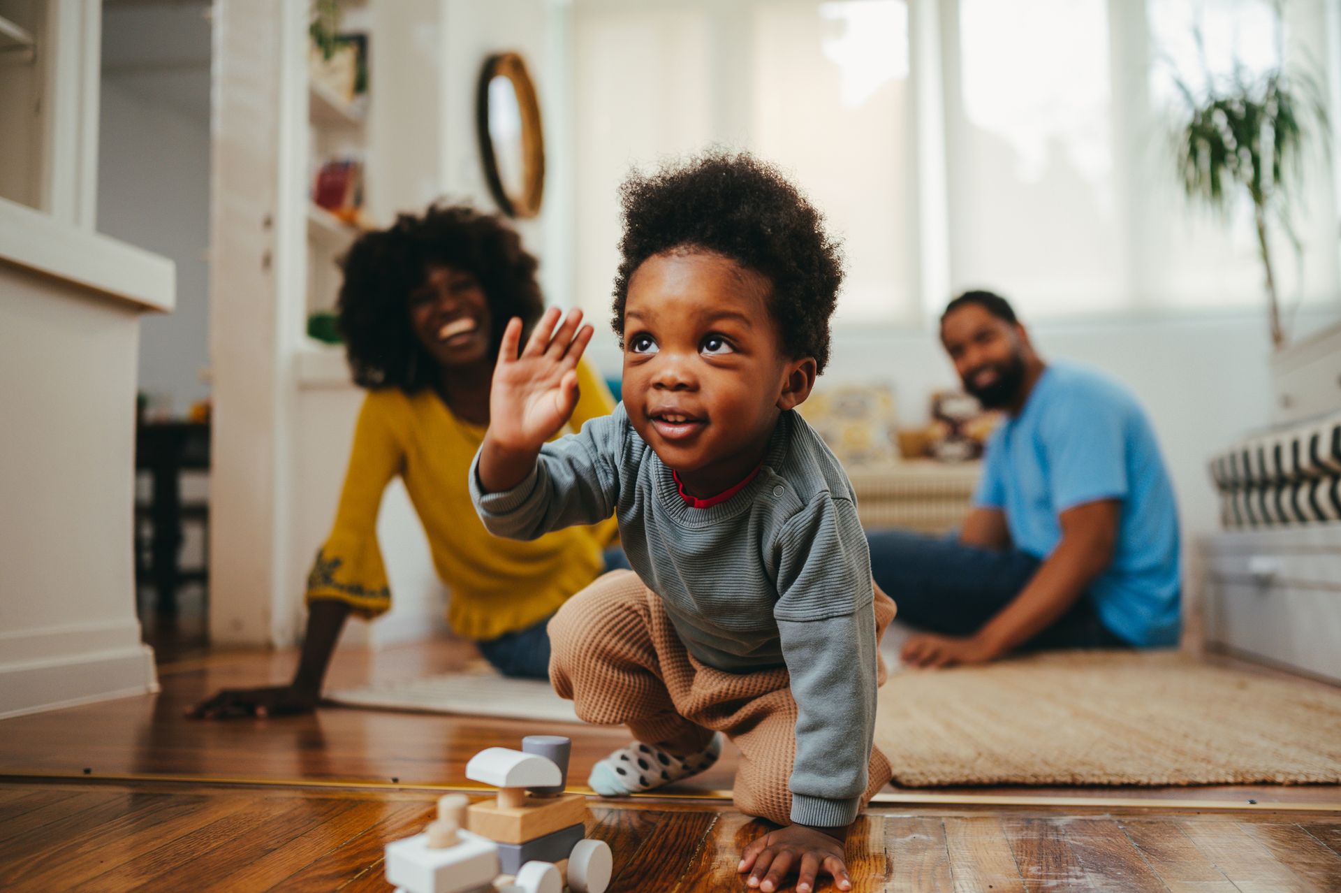 Young Black child waving, playing with toys on a wooden floor, with smiling parents in the background. Young Black child waving, playing with toys on a wooden floor, with smiling parents in the background.