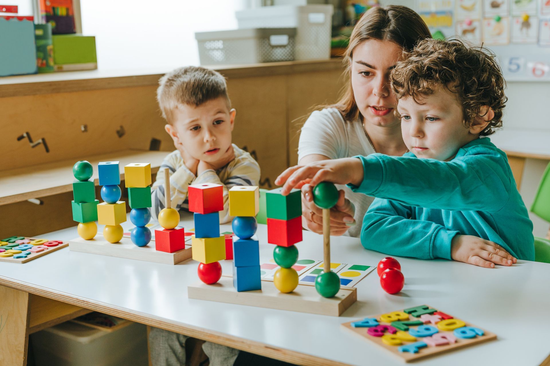 A teacher and two young boys play with colorful wooden blocks on a table, focused on stacking them.