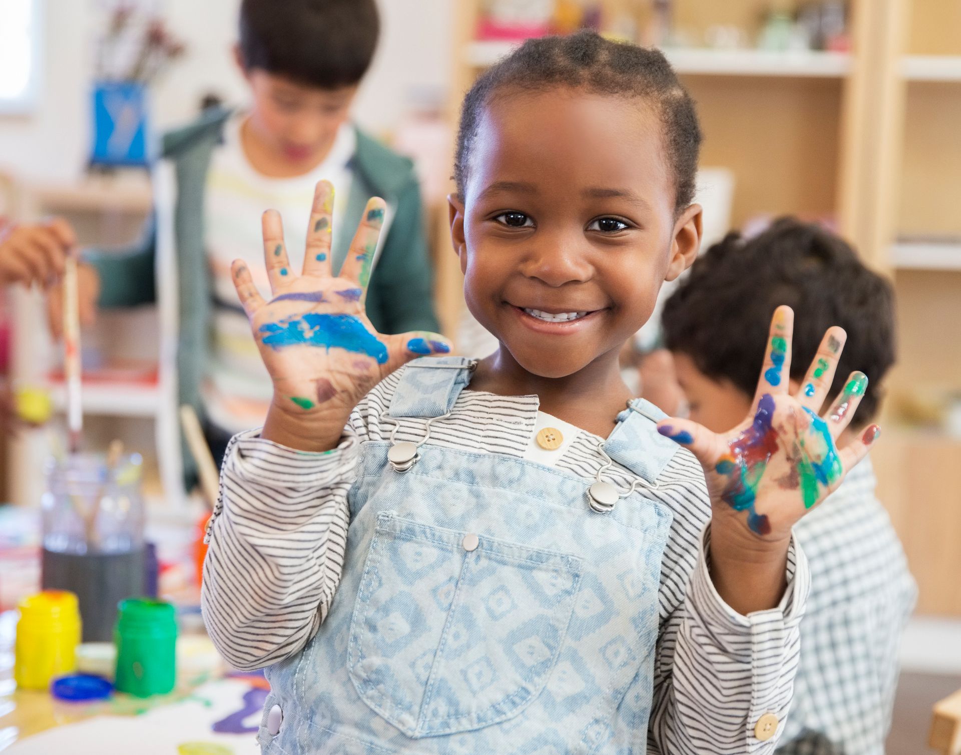 Smiling Black child with paint-covered hands in an art classroom. Other children and art supplies are visible in the background.