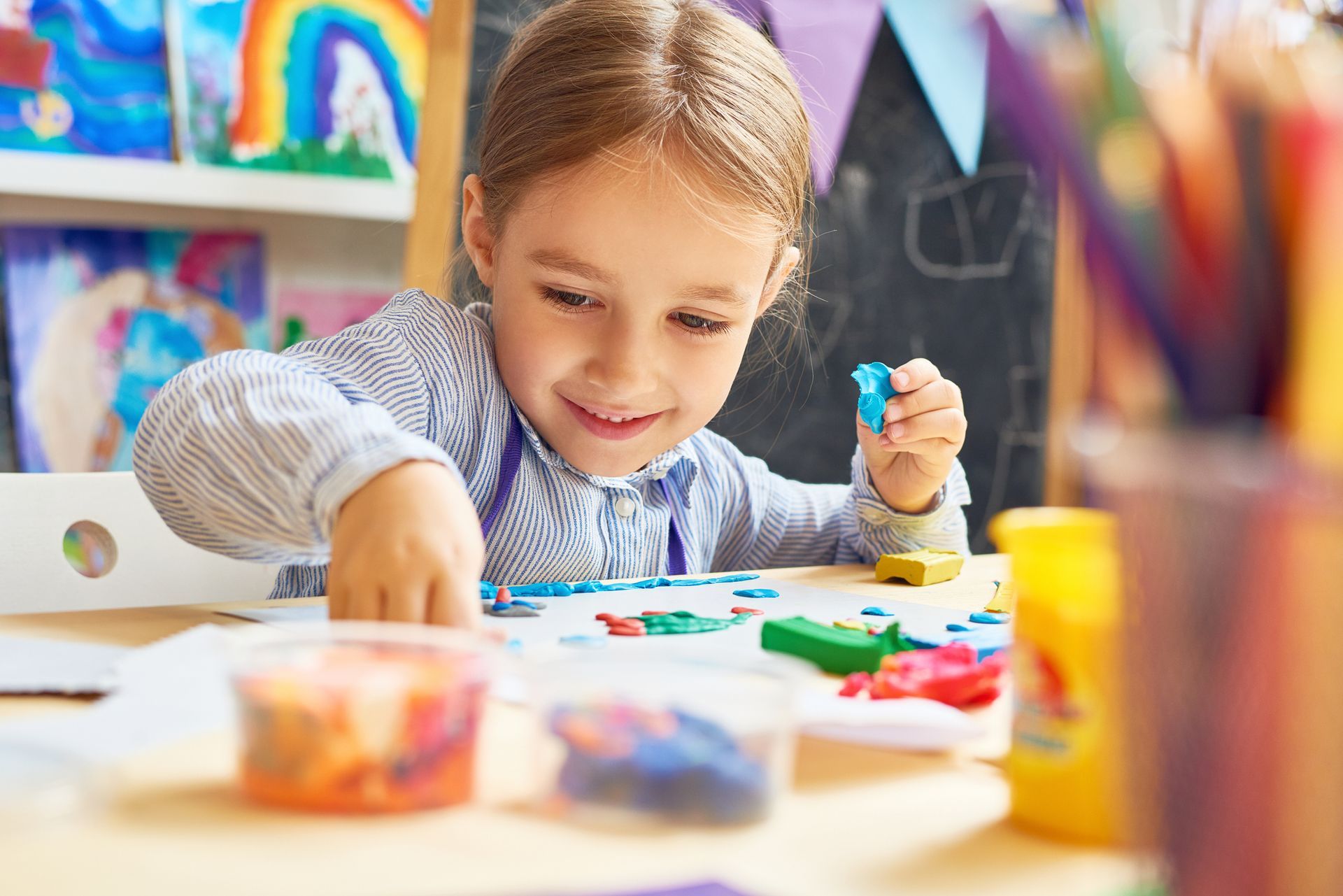 A young girl with a smile is playing with colorful clay at a table in an art studio, looking engaged and creative.