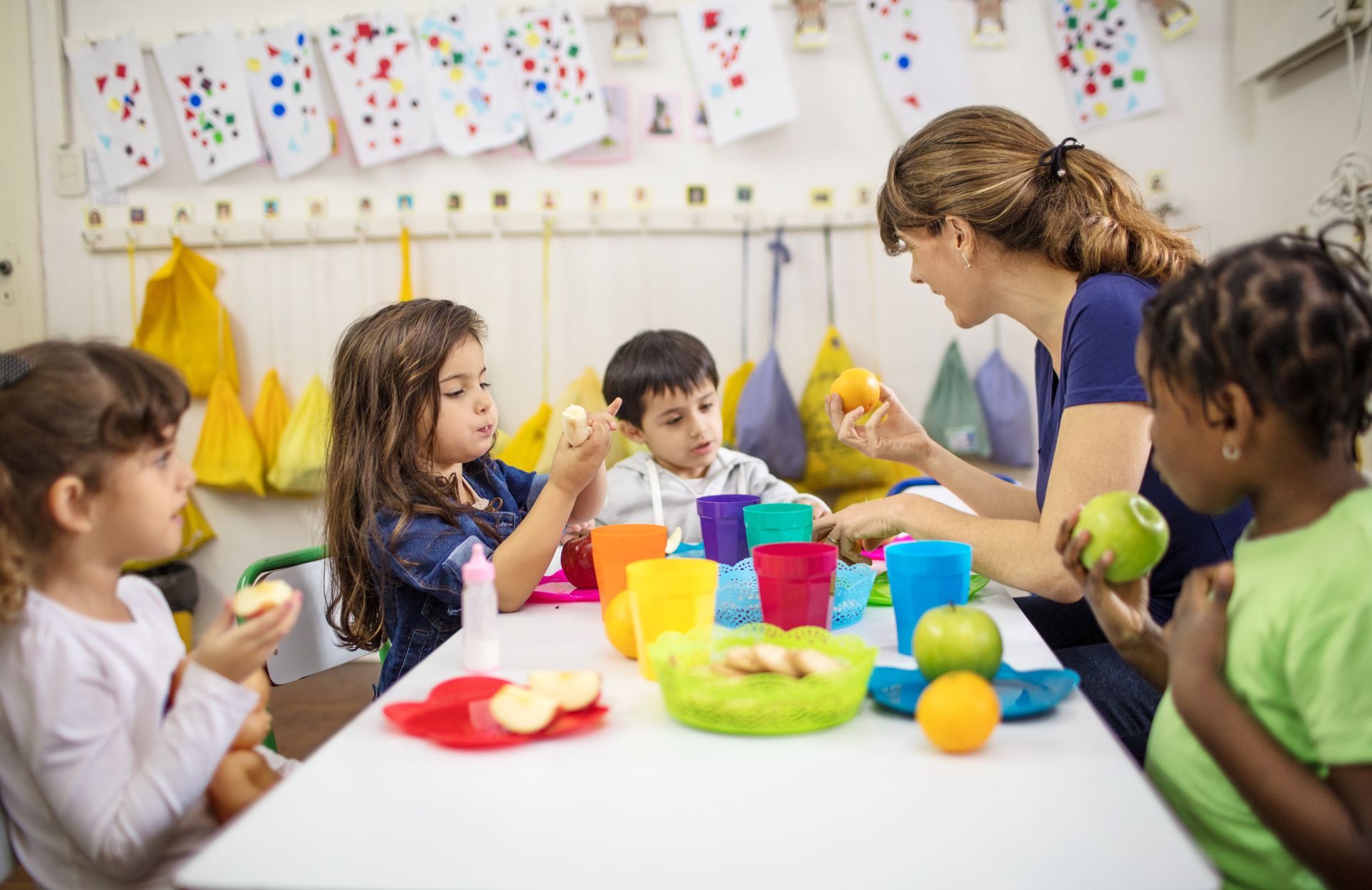 A teacher and four children at a table eating fruit in a colorful classroom. The teacher is smiling and talking.