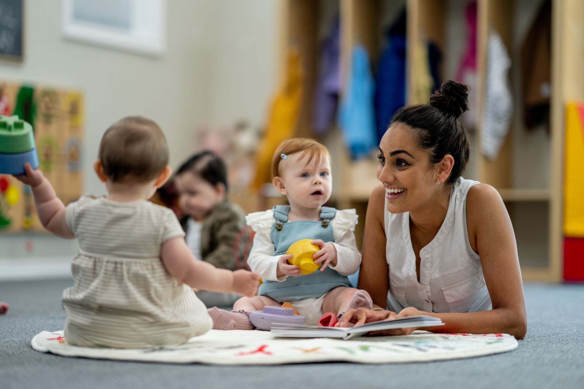 A daycare worker reads to three babies on a mat. The worker smiles while the babies look on, and one holds a toy.