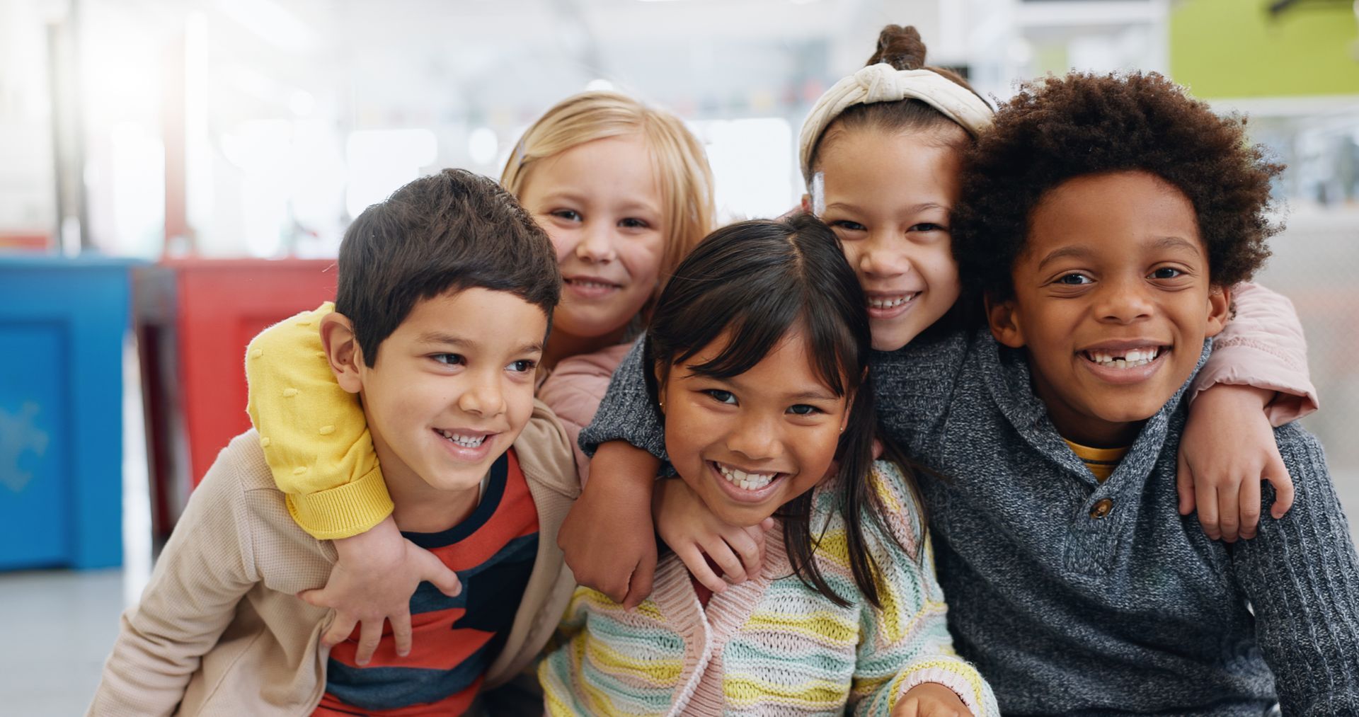 Group of five diverse children smiling and embracing, possibly in a classroom.