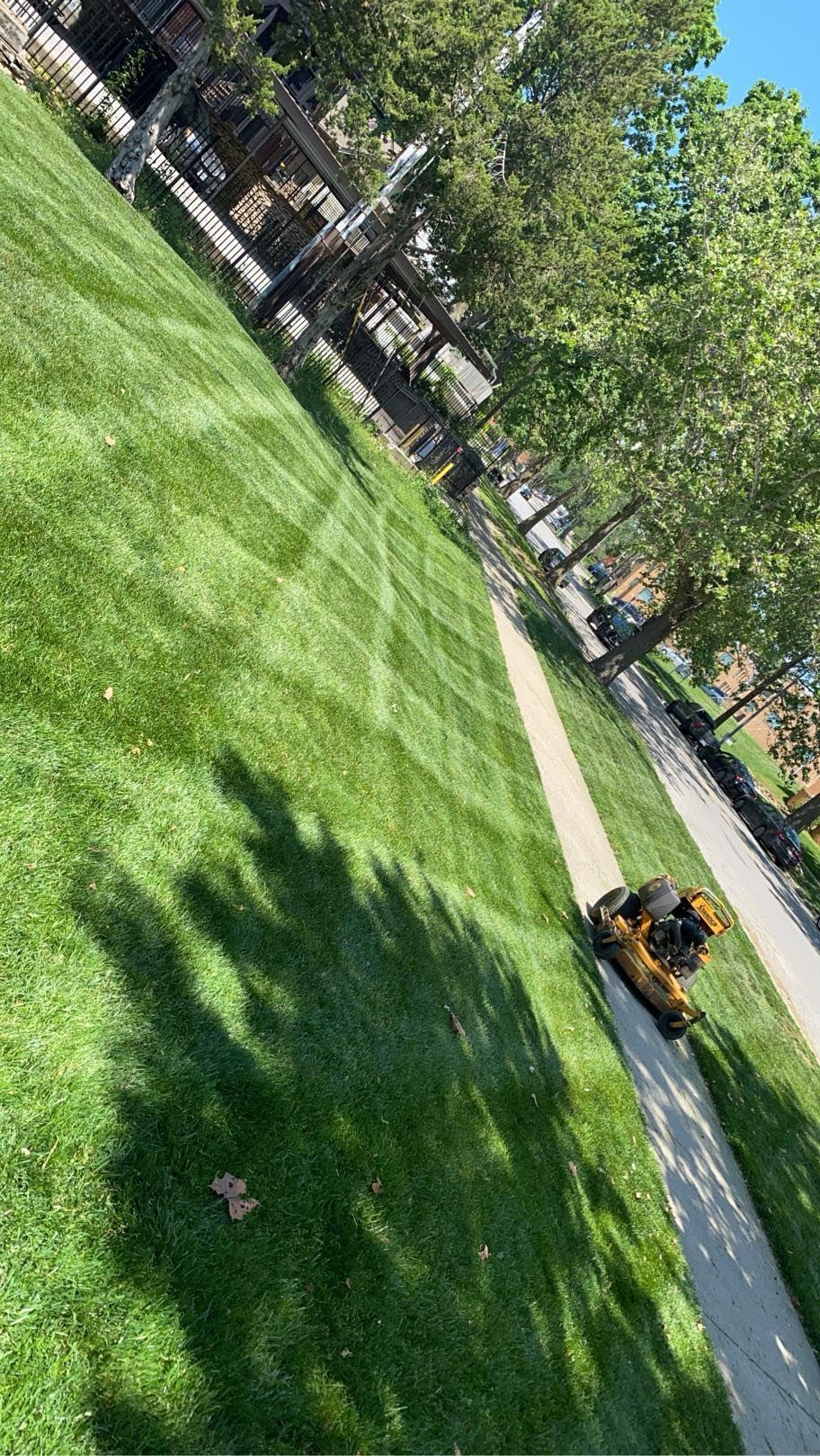 A person is mowing a lush green lawn next to a sidewalk.